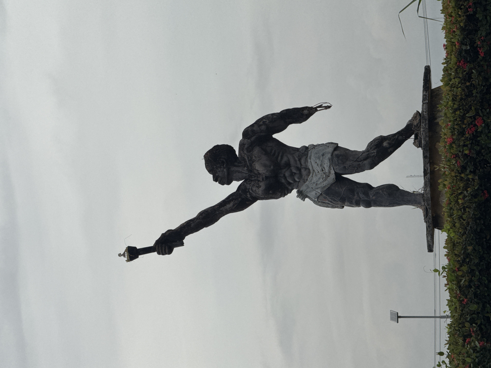 The image shows a statue of a muscular, dark-skinned man in Ihiagwa, Nigeria. He is depicted in mid-stride, appearing to leap forward with his right arm extended, holding an object resembling a torch or staff. He is wearing a cloth around his waist. The statue is mounted on a concrete platform covered with leafy plants and small red flowers. In the background, there is an overcast sky. A solar panel on a pole is visible in the lower right.
