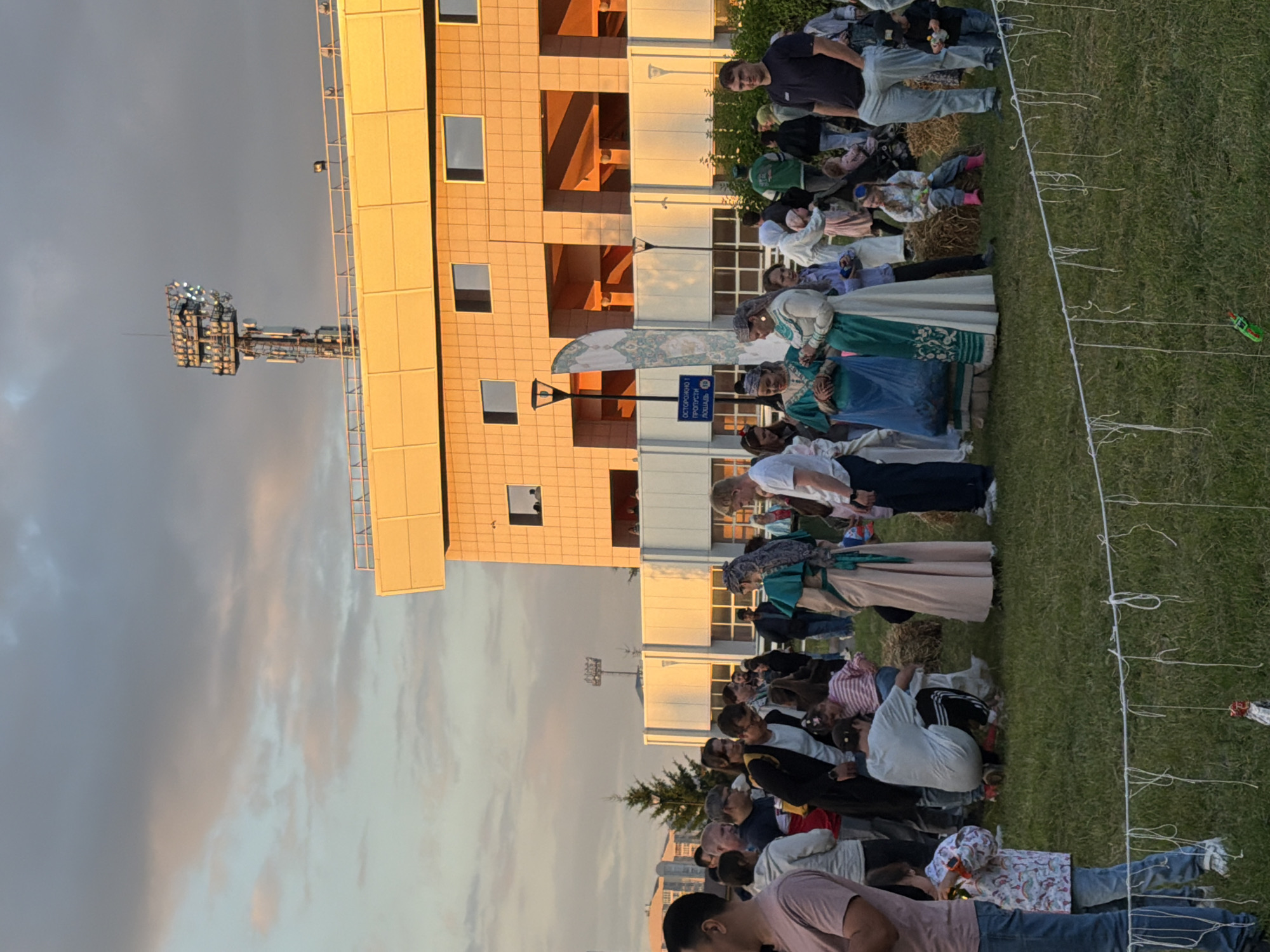 The image depicts an outdoor gathering in front of a modern building with a distinctive orange facade and white accents, likely a stadium or arena, given the floodlights visible at the top. The sky is a gradient of blue and pink, suggesting it is either sunrise or sunset. A diverse group of people, including adults and children, are milling around on a grassy area. Several individuals are dressed in traditional or cultural attire, with women in long, ornate dresses and headscarves. Others are in casual modern clothing. Some people are standing and talking, while others appear to be observing or participating in an event. There are bales of hay scattered on the grass, and a string barrier with hanging elements runs across the foreground, suggesting a delimited area or an activity. The overall atmosphere appears festive and communal.