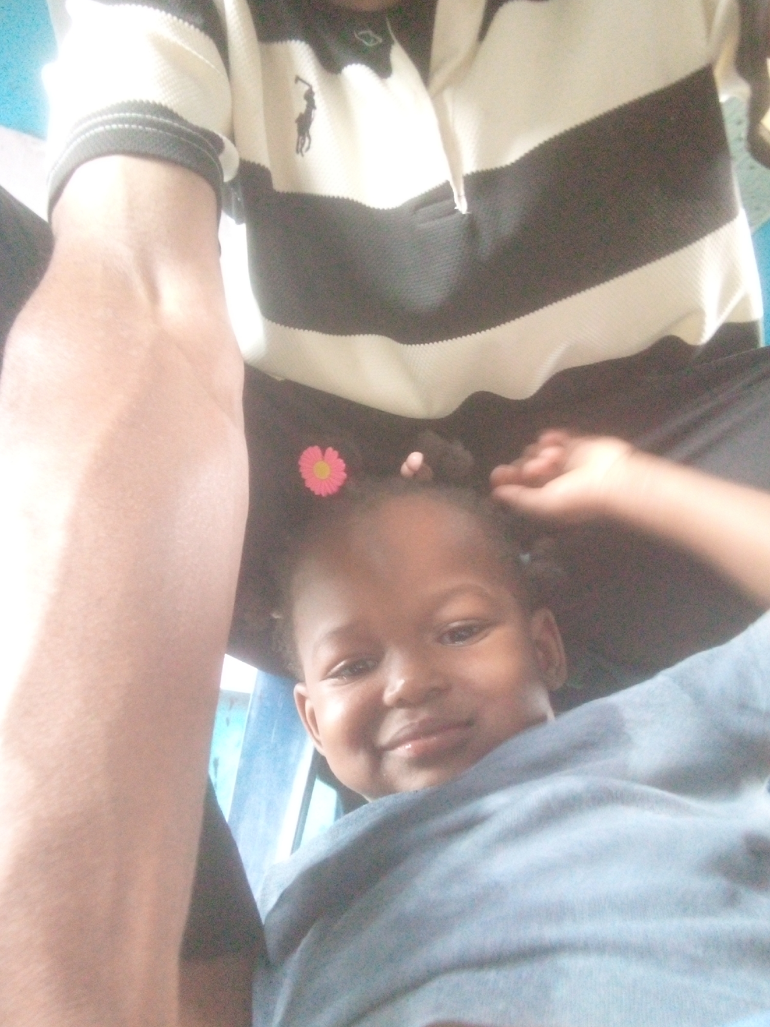A close-up indoor shot shows a small child with a pink flower hair accessory nestled under the arm of an adult. The adult is wearing a black and white striped polo shirt. The child is wearing a light blue shirt. The location appears to be in the city of Unknown, Unknown.