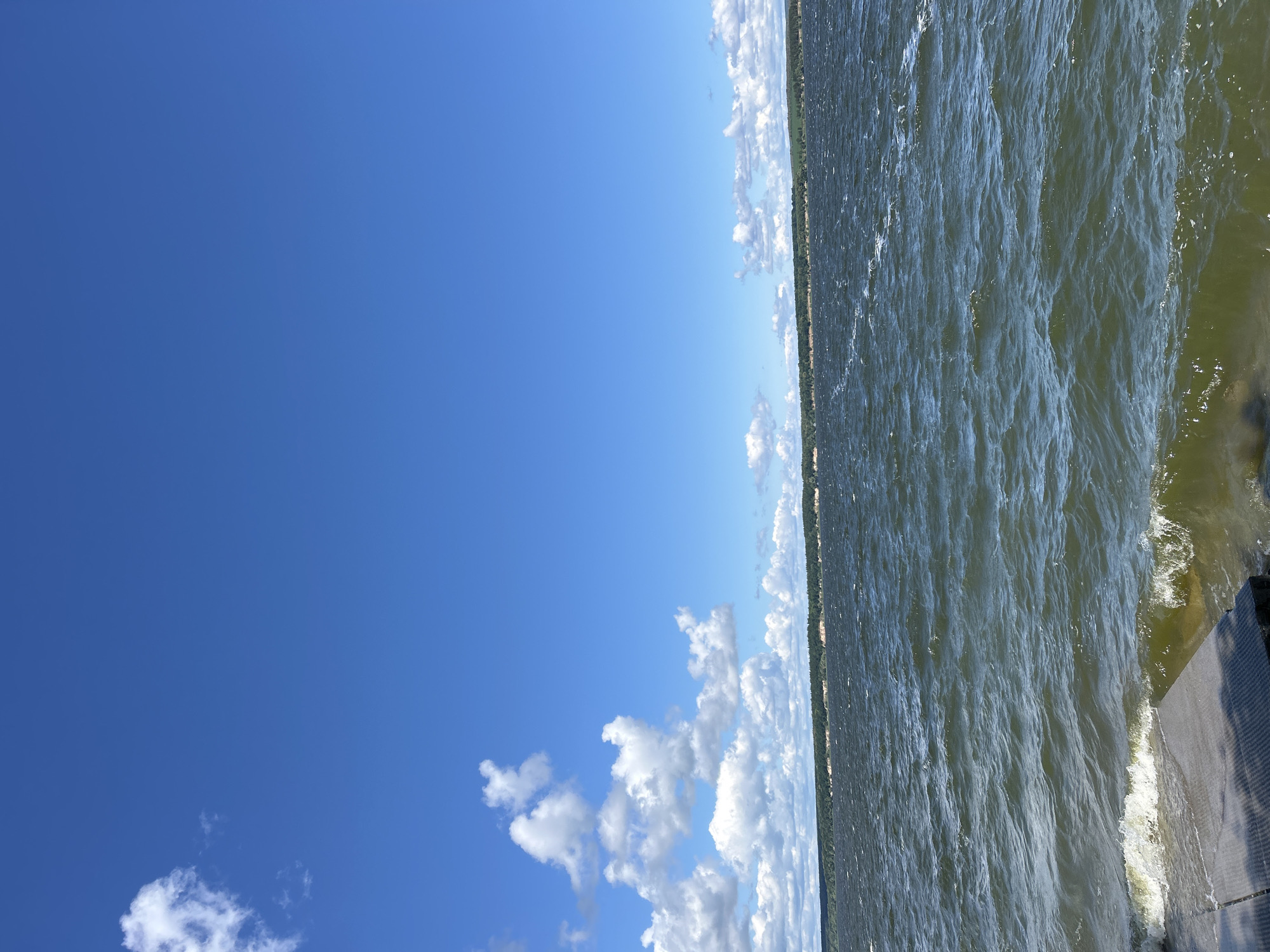The image displays a bright, sunny day with a clear blue sky. Fluffy white clouds are scattered across the sky, particularly on the left side of the frame. The right side of the image is dominated by a body of water, likely a lake or river, with gentle waves creating ripples on the surface. The water appears greenish-brown in color. Along the far horizon, a narrow strip of green land with trees is visible, suggesting a forested shoreline. In the bottom right corner, a portion of a dark, textured surface, possibly a dock or platform, is visible, with a shadow cast upon it. The scene evokes a sense of a peaceful outdoor environment. The time of day appears to be daytime, with the sunlight casting a bright glow. The weather is clear and likely warm. No people or text are visible in the image.