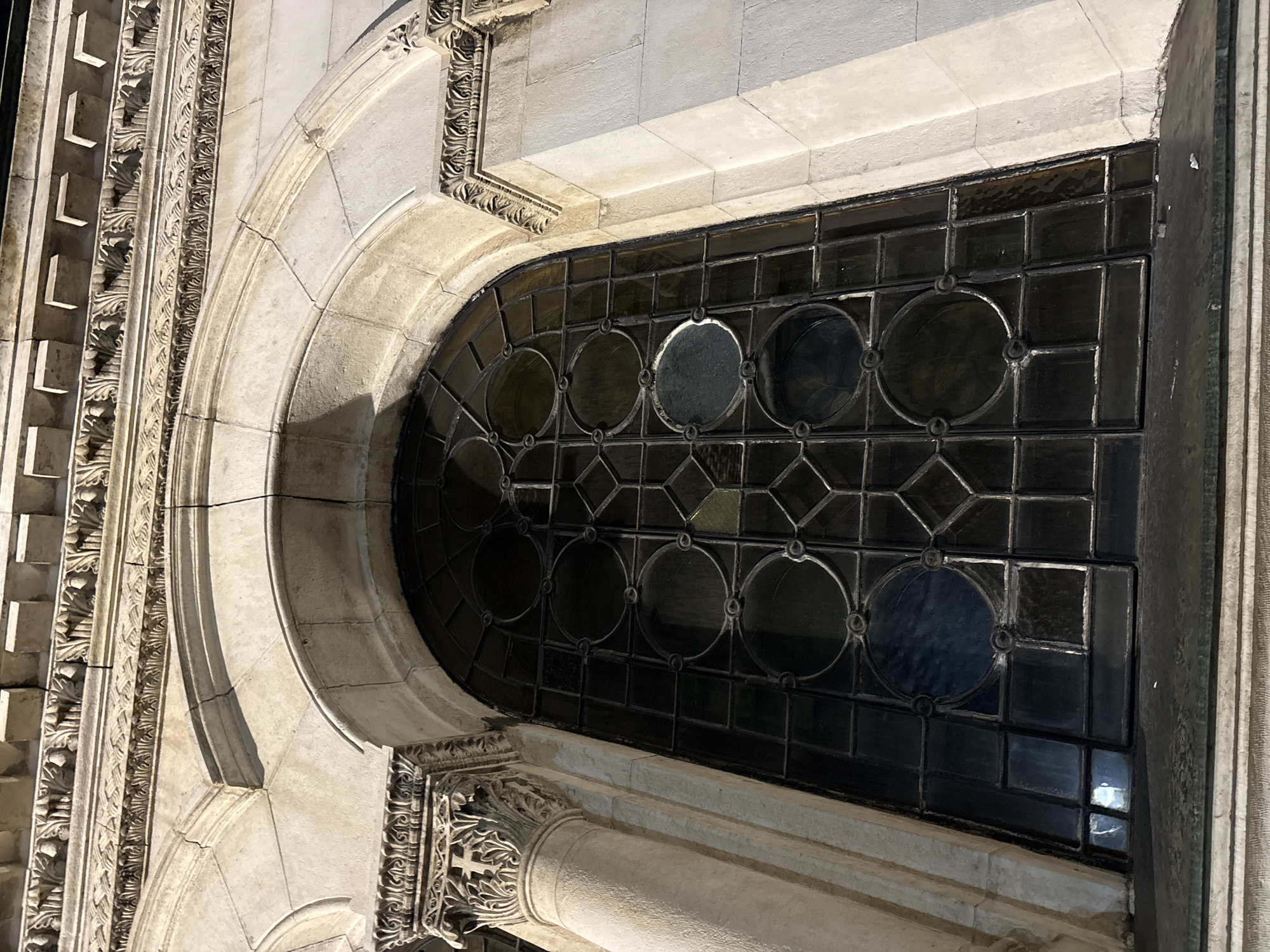 The media file presents a detailed close-up of a grand arched window, likely part of a historic building in Sofia, Bulgaria. The window itself features dark-toned, leaded glass, arranged in a complex geometric pattern. Several large circular panes are prominent, interspersed within a grid of smaller rectangular and diamond-shaped glass sections. Some of the circular panes show reflections or hints of a view, suggesting clear glass or subtle coloration.
The window is set within an elaborate stone facade, crafted from light-colored material, possibly limestone or marble. An ornate arch frames the window, adorned with intricate relief carvings along its curves. To the left of the window, vertical architectural elements are richly decorated with multiple layers of decorative motifs, including repetitive geometric patterns and detailed floral or acanthus-leaf designs. In the lower right, a finely carved column capital or pilaster with fluting and foliage patterns adds to the architectural grandeur.
The scene is bathed in bright daylight, casting distinct shadows that emphasize the texture and three-dimensional depth of the architectural carvings. This strong lighting suggests a clear, sunny day. No human activity or visible text is present, directing full attention to the exquisite craftsmanship and historical aesthetic of the architecture. The overall impression is one of significant historical artistry and detailed construction.
