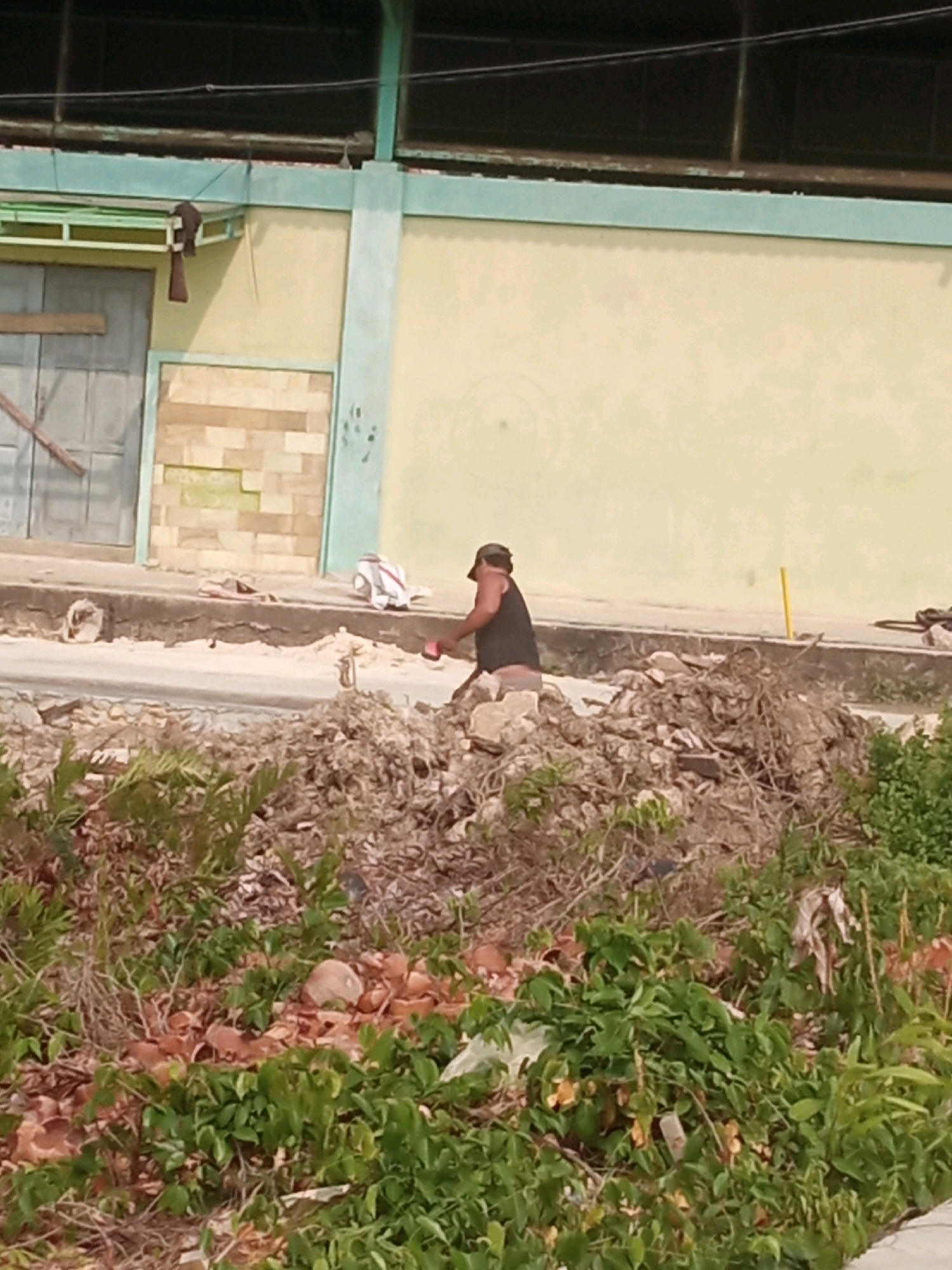 A man wearing a dark tank top and a cap is seen from the side, bending over to pick up debris in a pile of rubble. He is right-handed and is holding a red object, possibly a shovel, in his right hand. The debris consists of rocks, broken concrete, branches, and various forms of waste. In the background, there is a light-colored building with a teal-colored roofline and trim. A large, gray door with wooden planks is visible to the left of the building. The ground in front of the building is a paved walkway with some scattered debris. The scene appears to be an outdoor area undergoing cleanup or demolition in an urban environment. This scene is located in the city of Bontang, Indonesia.
