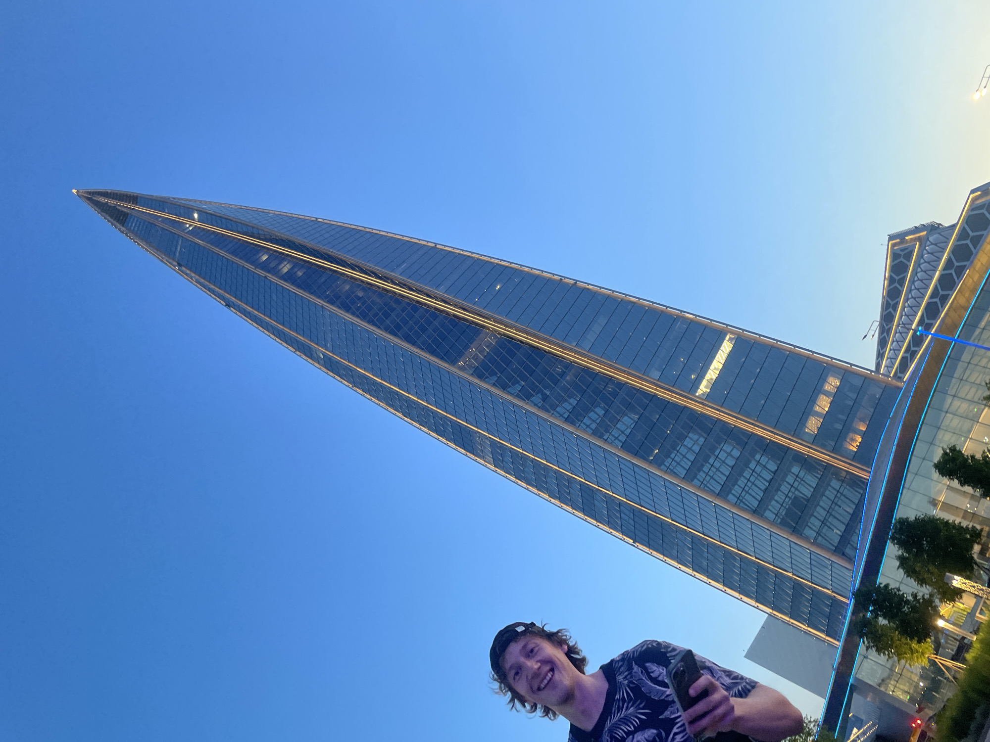 A man wearing a black t-shirt with a tropical leaf pattern and a black baseball cap smiles at the camera, holding a smartphone in his left hand. He is positioned on the left side of the frame, looking slightly upward. Behind him, a very tall, slender skyscraper with a pointed apex dominates the right side of the image. The skyscraper's facade is made of glass and is illuminated by warm yellow lights along its edges. The sky is a clear, bright blue. In the foreground, at the base of the skyscraper, there are some trees and a building with a decorative facade featuring blue lighting. The overall scene suggests an outdoor setting in a modern urban environment, possibly during the evening or early morning due to the lighting. The location is identified as Unknown, Unknown.