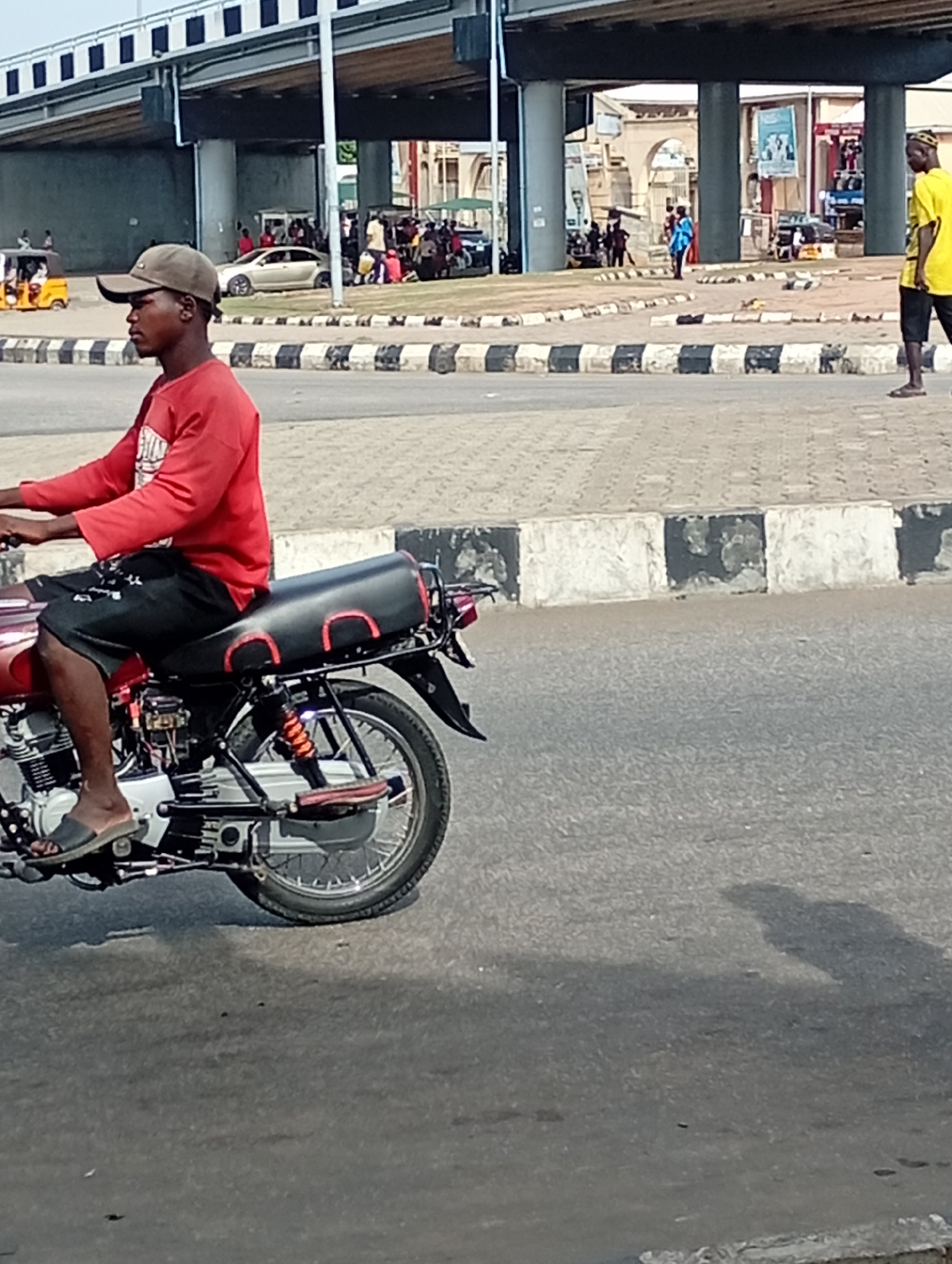 The image depicts a man riding a motorcycle on a street in Lokoja, Nigeria. The man is wearing a red long-sleeved shirt, black shorts, a baseball cap, and sandals. The motorcycle is red with a black seat. In the background, there is a road overpass, people, cars, and commercial tricycles. The curb separating the road from the pedestrian area is painted with black and white stripes. The ground is asphalt. Another man in a yellow shirt is visible walking in the background on the right side of the image.