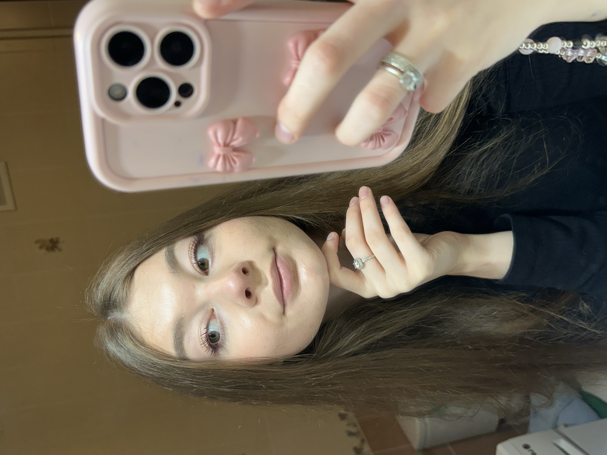 A young woman is captured in a mirror selfie, with the image rotated 90 degrees clockwise. She has long, straight, medium-brown hair, parted centrally, and light skin. Her large, light brown eyes are open, and she maintains a neutral expression with slightly parted, naturally full lips, enhanced by subtle makeup and mascara.
She holds a light pink smartphone, which features a distinct three-lens camera system and a flash. The phone is encased in a matching light pink cover, decorated with small, textured bow-shaped embellishments on its back. On the hand holding the phone, two silver-colored rings are visible. Her other hand rests near her face, showcasing a prominent silver-colored ring with a clear gemstone on her ring finger. A beaded accessory, comprising light pink and silver/grey beads, is partially visible around her neck or shoulder, worn over a black top.
The background is a soft, out-of-focus, light brown or beige wall, suggesting an indoor setting. The lighting is even and soft, casting a gentle sheen on her skin. The photo is a direct reflection, taken as a mirror selfie in what appears to be a casual indoor environment in Kazan, Russia. There is no visible text within the image.