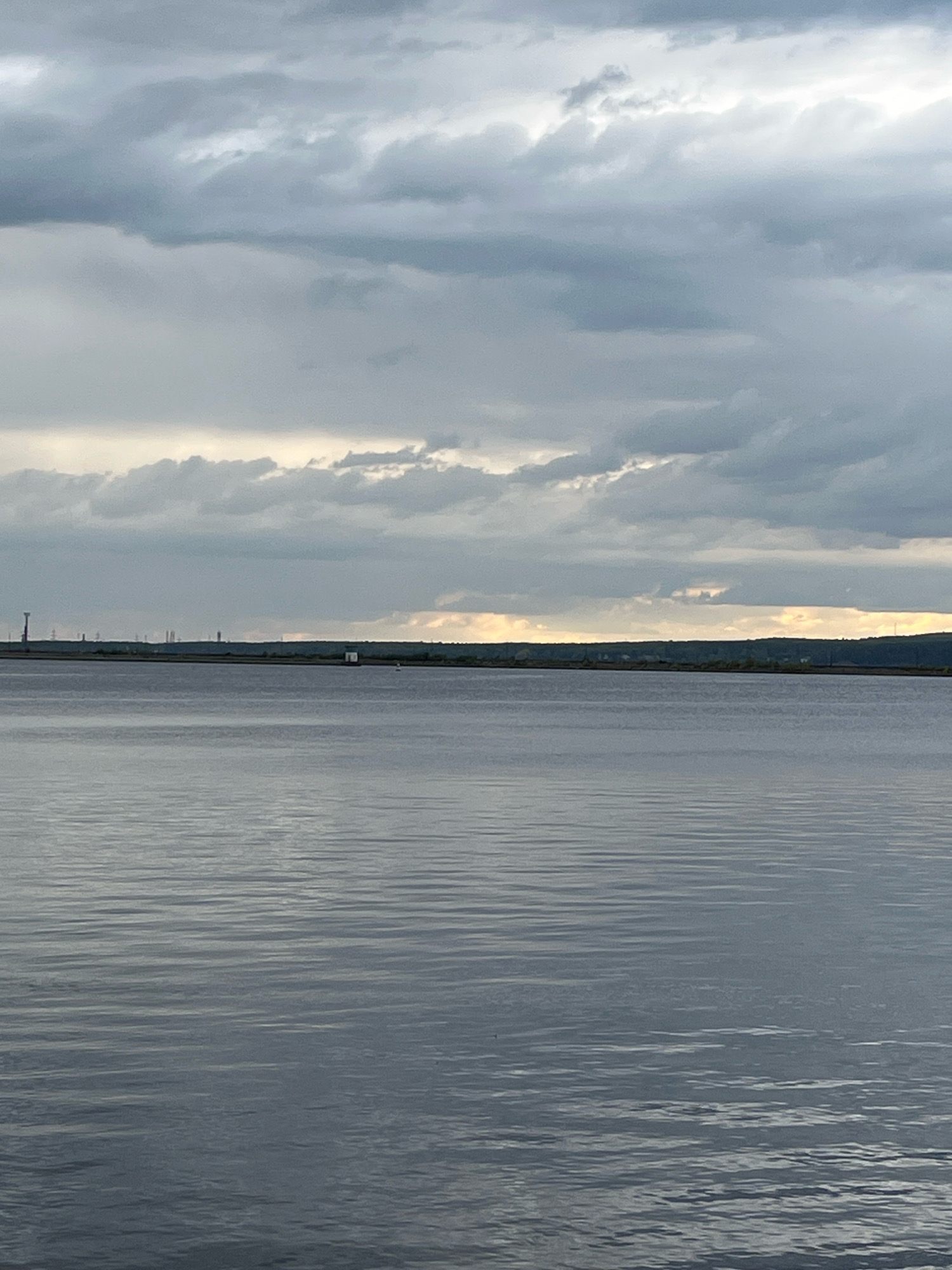 The image depicts a calm body of water with a distant, tree-lined shore under a cloudy sky. In the distance, there are several industrial structures, including tall towers, and a small white building on the shore. The water's surface is gently rippled, reflecting the muted light. The scene appears to be a natural landscape with some man-made elements on the horizon. The location is the Kama River in Chaikovsky, Russia, as indicated by the presence of industrial facilities typical of the area. There are no people visible in the image.