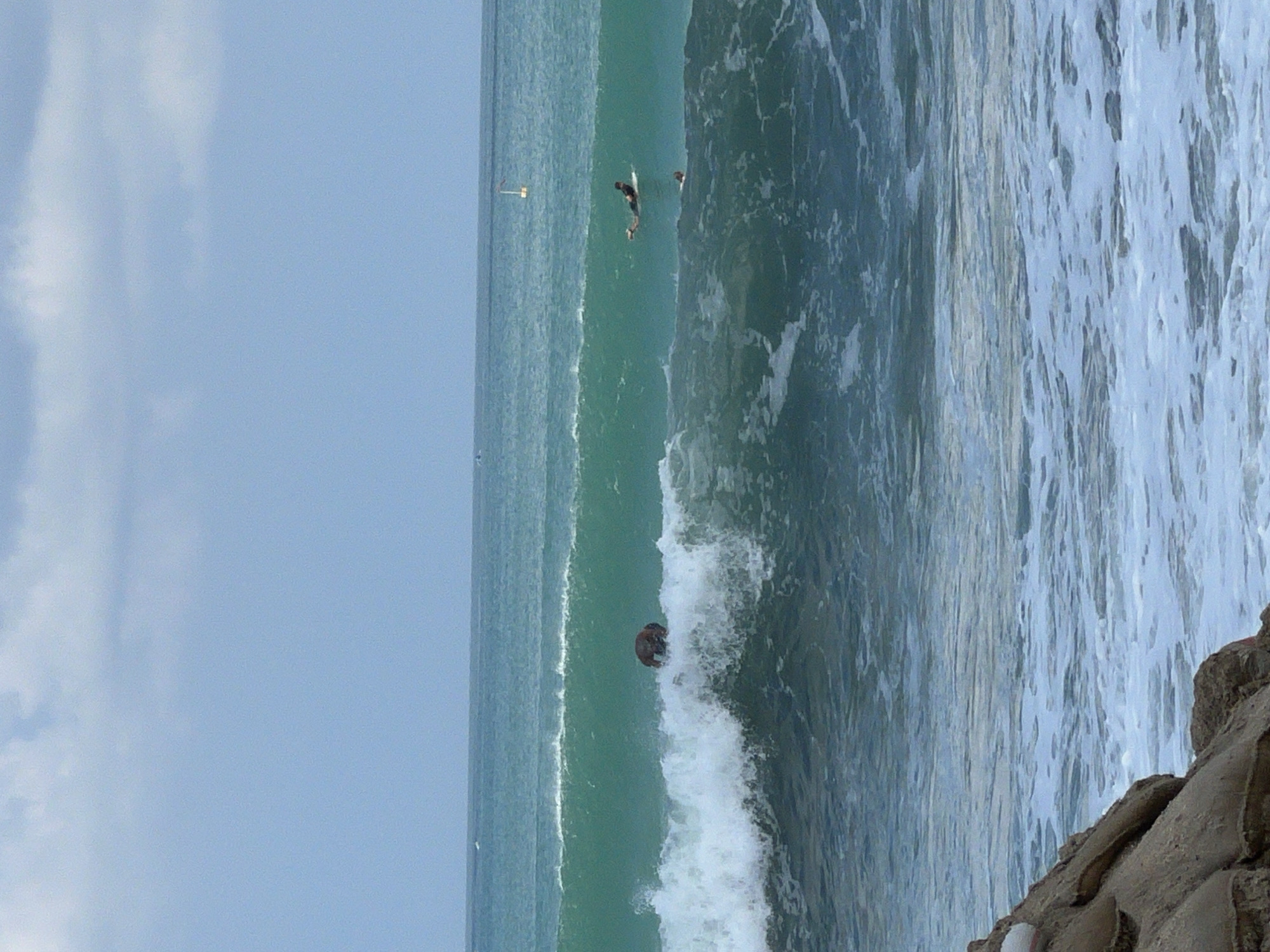 The image depicts a medium shot of the ocean from the perspective of a beach. In the foreground, rough, brown rocks are visible on the left, forming part of the coastline. The middle ground is dominated by a large, turquoise wave that is breaking. A person on a surfboard is positioned in the trough of this wave, appearing to be either paddling or waiting for a ride. Further out, the ocean is a deeper blue, with several more surfers visible on their boards. In the background, a buoy with a yellow flag can be seen in the distance. The sky is overcast with light gray clouds. The scene appears to be a typical surfing spot.