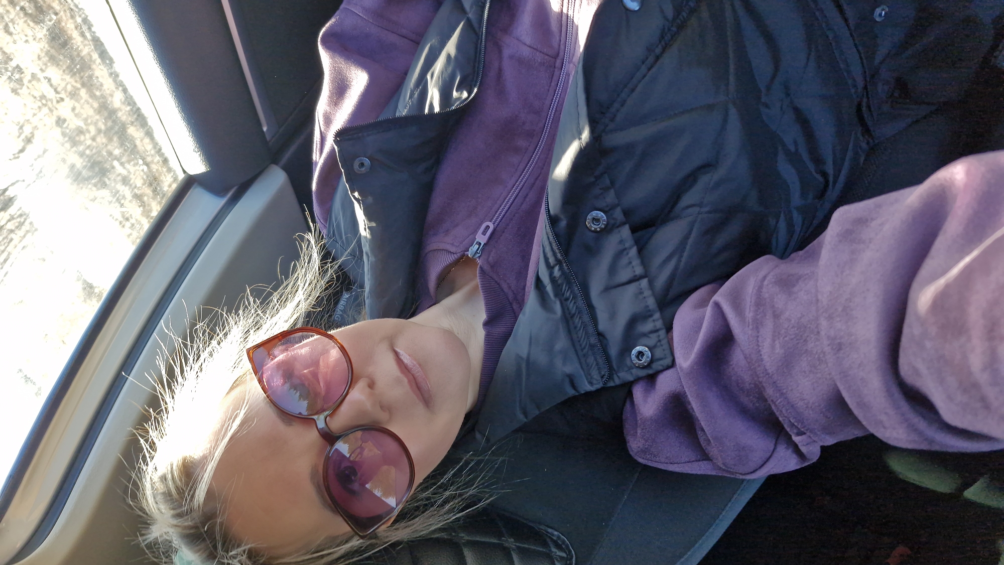 A person, likely female, with light-colored hair and wearing sunglasses, is depicted resting or relaxing inside a vehicle. The image is taken from a high angle, with the subject oriented horizontally across the frame. She wears a purple zippered hoodie, partially open to reveal a hint of skin at the neck, layered under a black quilted puffer vest with snap buttons.
Bright sunlight illuminates her blonde hair, creating a halo effect, and highlights the side of her face. She wears sunglasses with reddish-brown frames and pinkish, reflective lenses that obscure her eyes. Her mouth is slightly ajar, suggesting a relaxed state. Behind her head and shoulder, the word "AIRBAG" is visible in white lettering on a dark panel, indicating a car or vehicle interior.
To the left of the frame, through the vehicle's window, a very bright, out-of-focus exterior landscape is visible, suggesting a sunny daytime setting. The overall impression is one of calm or repose during travel in what appears to be a cool but bright environment, possibly within Otchor, Russia.