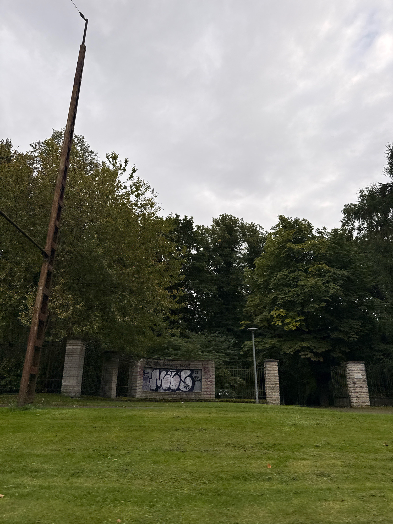 The image depicts a gray, overcast sky over a grassy field with a row of trees in the background. In the middle ground, a brick wall with graffiti is visible, flanked by stone pillars and metal gates. To the left, a tall, rusty metal pole stands prominently. The scene appears to be an entrance to a park or an abandoned area. The graffiti on the wall suggests it is located in an urban environment, possibly a public space. The overall impression is one of neglect or disuse.