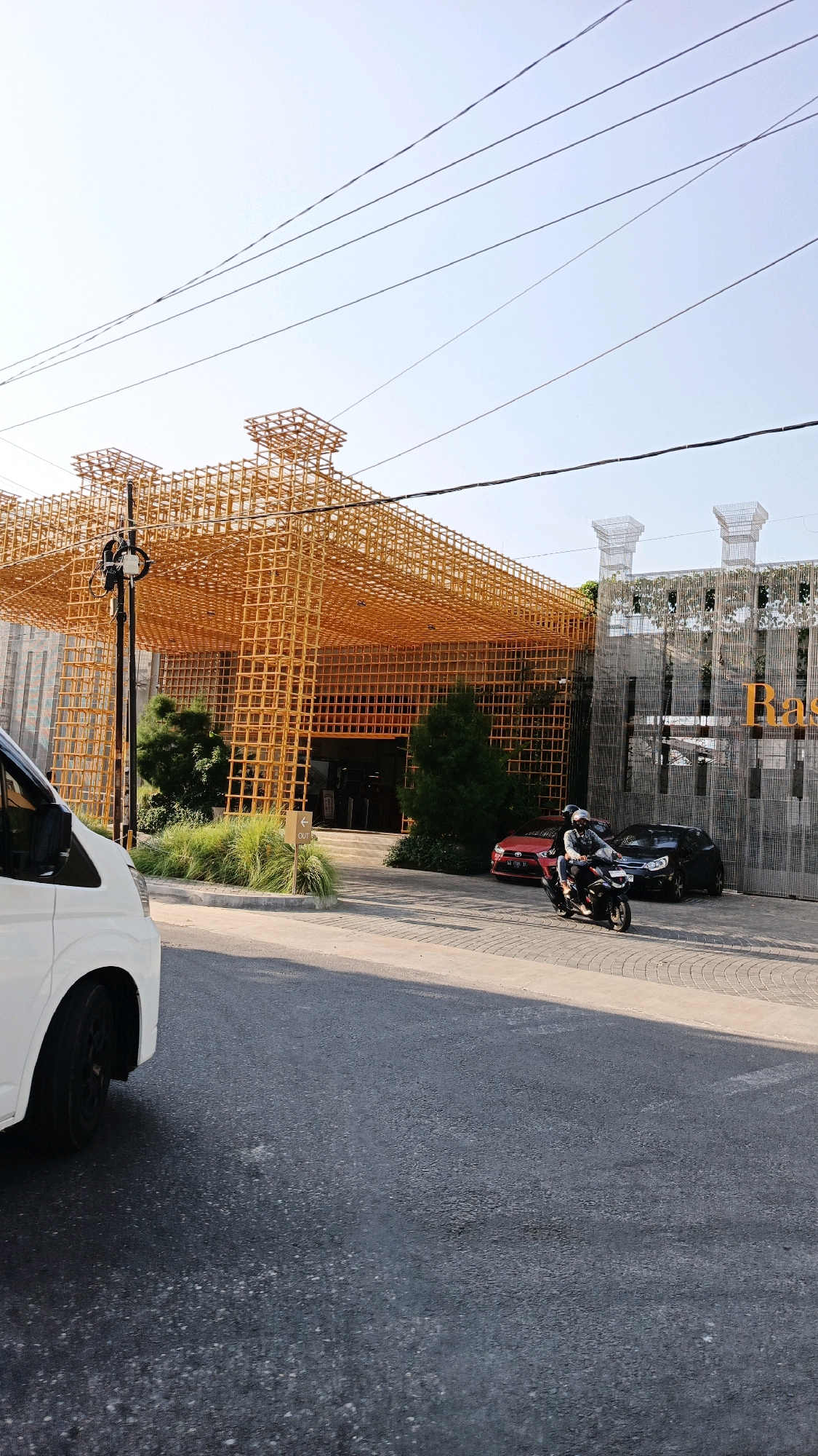 A street-level, wide shot captures the exterior of a building with a distinctive orange lattice structure covering its facade. The building appears to be a commercial establishment, possibly a restaurant or shop, with large glass doors visible at the entrance. In front of the building, a paved area serves as a parking lot and walkway.
In the foreground, the left side of a white van is visible, its front wheel turned slightly. To the right of the van, a motorcycle carrying two individuals, a driver and a passenger, is moving away from the viewer. The driver is wearing a helmet and a light-colored jacket, while the passenger is also wearing a helmet.
Behind the motorcycle, a red sedan is parked. Further to the right, a black sedan is parked. Lush green trees and ornamental grasses are planted around the building's entrance. Overhead, power lines crisscross the sky, which is a pale blue with a slight haze. The lighting suggests it is daytime, with the sun casting shadows.
The building's facade is a prominent feature, composed of interconnected orange rectangular elements creating a voluminous, grid-like shell. The name "Ras" is visible in large yellow lettering on the right side of the building's facade, suggesting it may be called "Ras Restaurant" or a similar establishment. The architectural style appears modern and bold. The road in the foreground is asphalt. The overall setting appears to be a public street in a developed area, likely in a city. Given the context provided, this scene is located in Ngadirejo, Indonesia.
