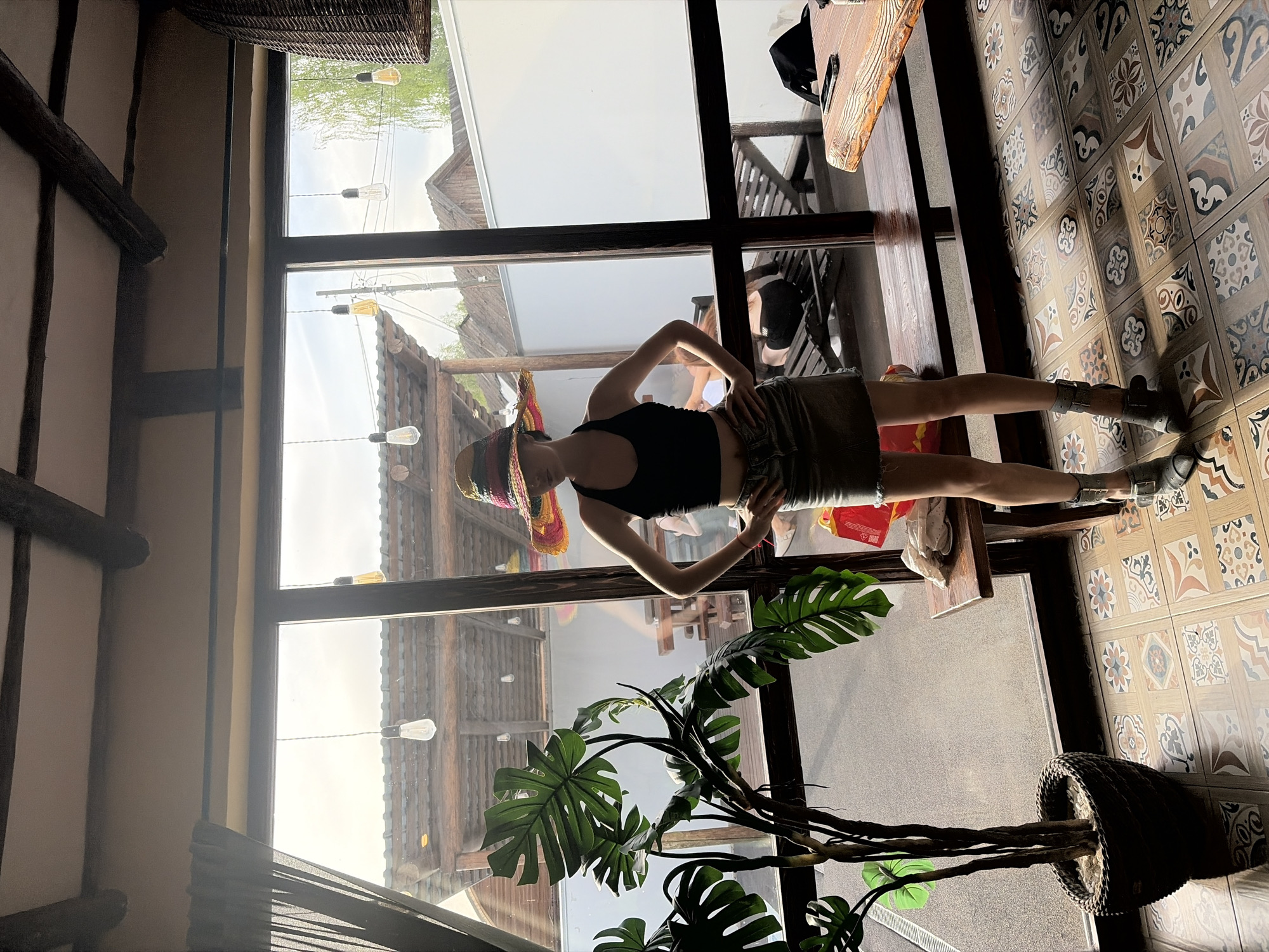 The image shows a woman indoors, wearing a black top, khaki skirt, and a colorful sombrero. She is standing with her hands on her hips, posing for the camera. The location appears to be a cafe or restaurant in Старое Михайловское, Russia, based on the rustic interior design including exposed wooden beams, a large window overlooking a building with similar architectural style, and decorative light bulbs strung outside. Inside, there's a large potted monstera plant in the foreground, a table and chairs in the background, and patterned floor tiles.