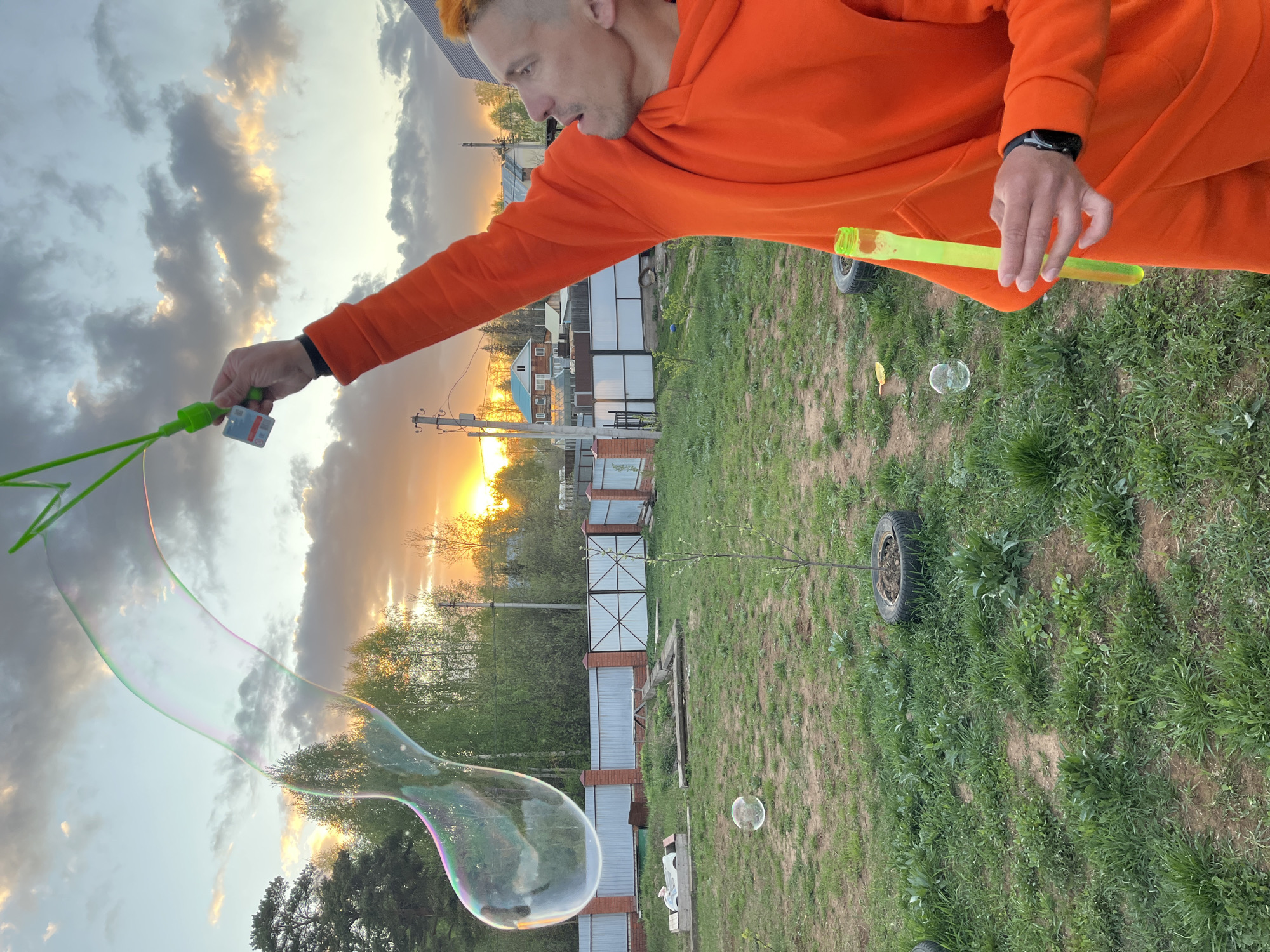A man in an orange hoodie blows bubbles in a grassy area, possibly in Chaikovsky, Russia, given the context. He holds a green bubble wand in his outstretched left hand and a yellow bubble solution container in his right hand. Several bubbles are visible, one large and teardrop-shaped, and others smaller, floating in the air. The background includes a wooden fence, trees, and buildings under a sky with clouds and a bright sunset. A tire lies on the grass in the mid-ground.