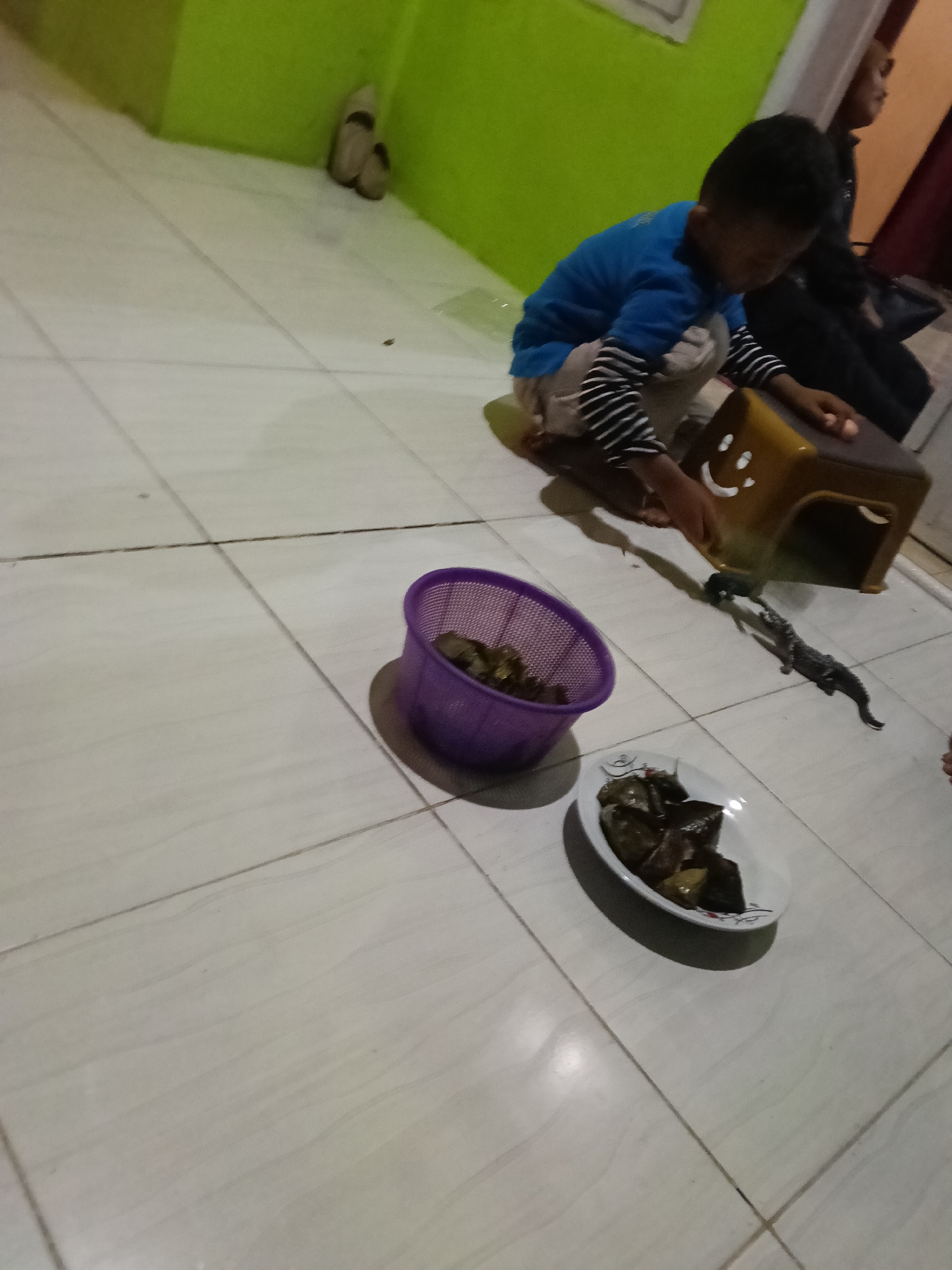 In an indoor setting with lime green walls and beige tiled flooring, a young boy wearing a blue shirt with black and white striped sleeves and light-colored pants kneels on the floor. He is interacting with a small, yellow plastic stool that has a smiley face drawn on it. In front of him, to his left, is a purple basket filled with dark green leafy items. To his right, on a white oval plate, is a pile of dark brown food items, possibly wrapped in leaves. Behind the boy, a woman wearing a hijab and dark clothing is partially visible, looking away from the camera. Further in the background, on the floor near the green wall, are two beige colored slippers. A dark, elongated object, possibly a toy or a prop, is situated between the basket and the plate. The lighting appears to be artificial, casting shadows on the tiled floor. The scene is likely a home environment in Paojan Lor, Indonesia, given the description of the location.