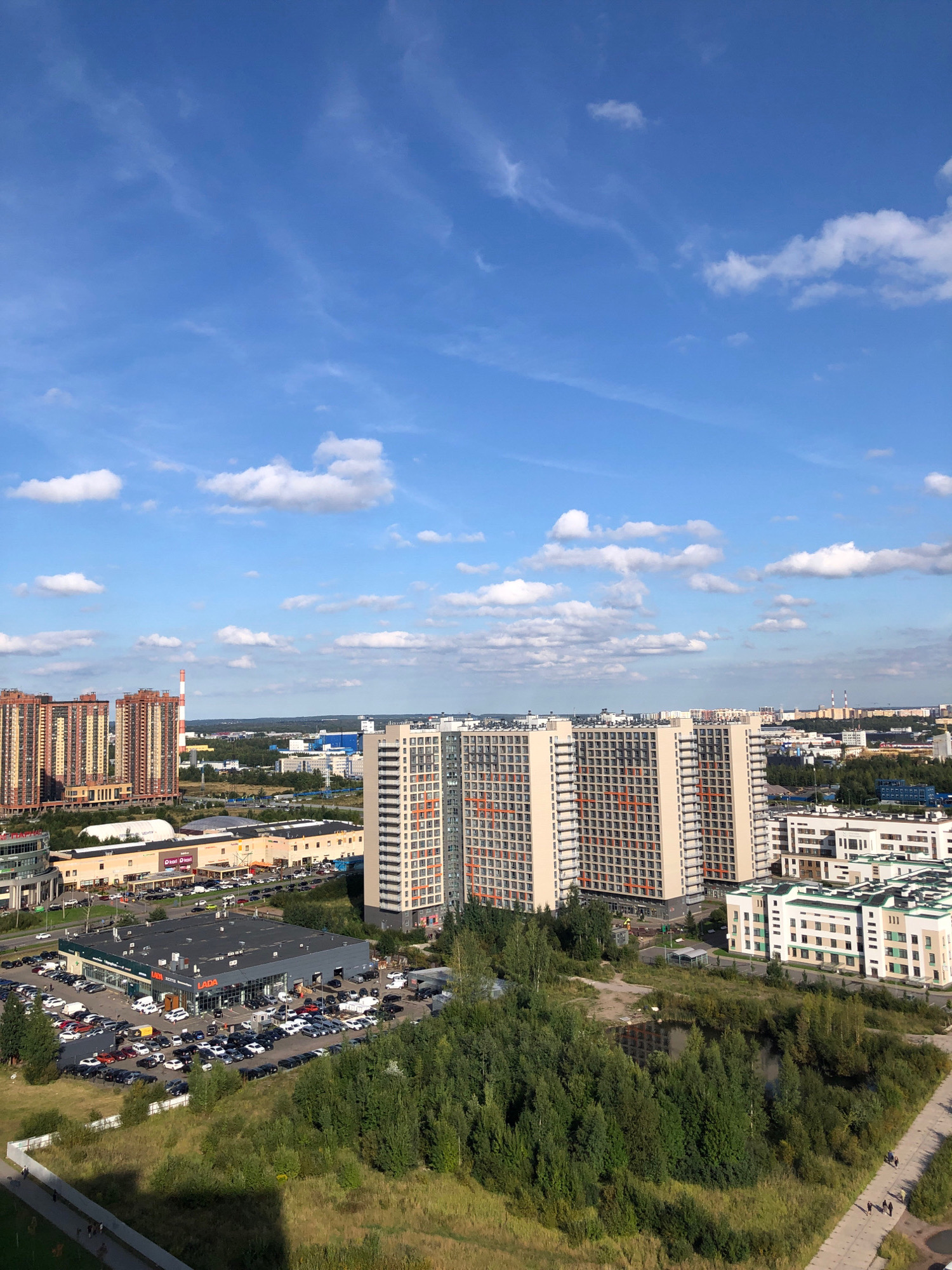 The image depicts a panoramic view of the city of Lavriki, Russia, under a bright blue sky with scattered white clouds. In the foreground, a large parking lot is filled with numerous cars, parked in front of a commercial building with "LADA" signage prominently displayed. Adjacent to this is a dense green forest. Further in the background, several modern high-rise apartment buildings dominate the skyline. A tall industrial chimney with red and white stripes is visible in the distance. The scene suggests a developing urban area with a mix of residential, commercial, and natural elements. Several people are walking on a paved path in the lower right corner of the image.