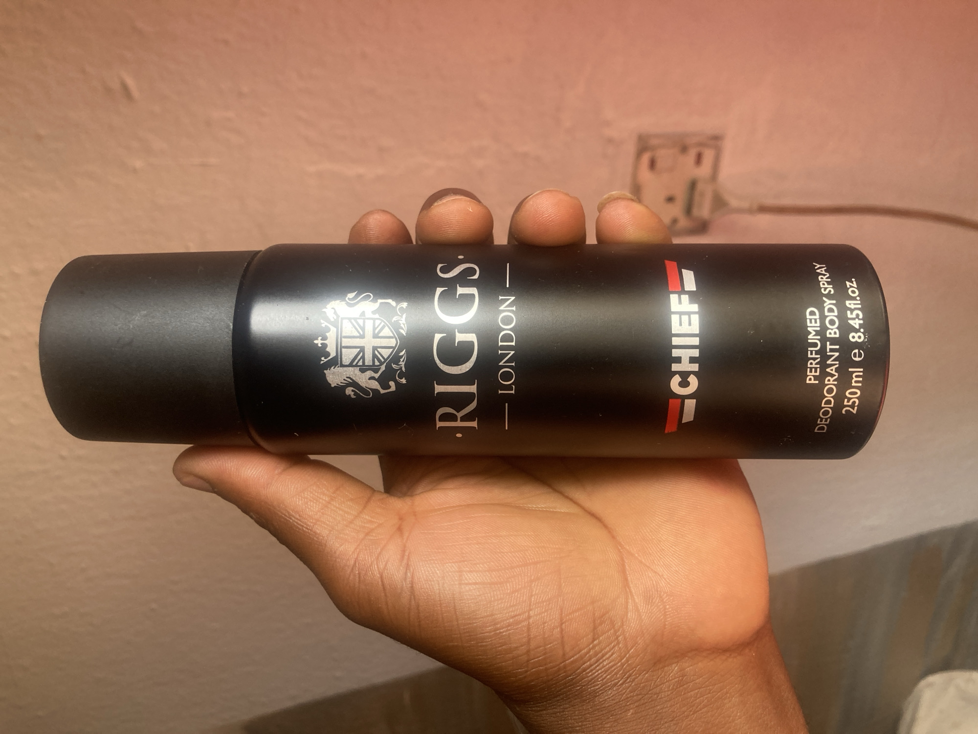 A black spray deodorant with the label "RIGGS LONDON - CHIEF - PERFUMED DEODORANT BODY SPRAY - 250 ml e 8.45fl.oz." is held in a person's hand. The background is a light pink wall and a white electrical outlet. The location is not discernible from the information in the image.