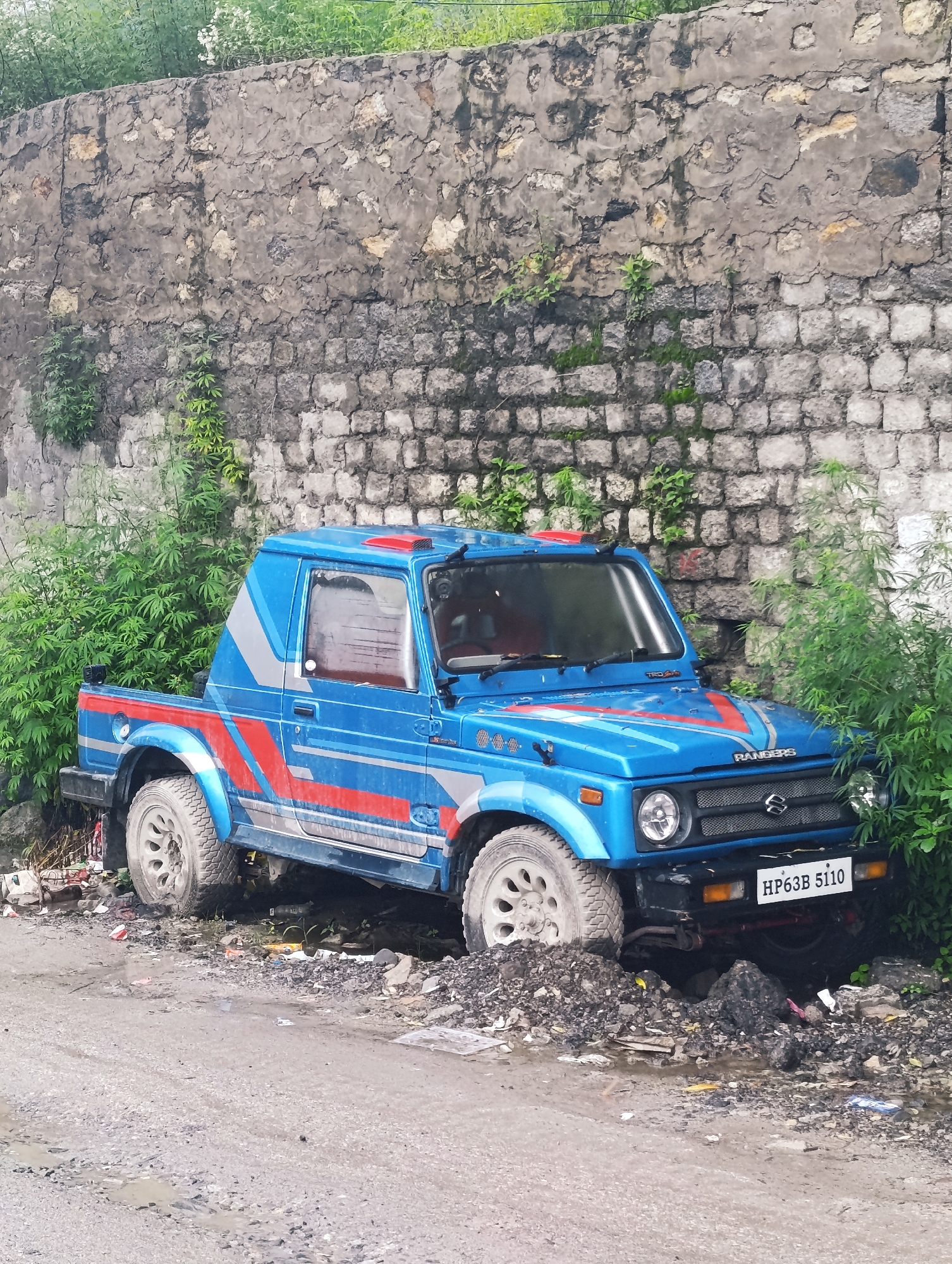 An eye-level, full shot captures a vibrant blue Suzuki Samurai pickup truck parked on a dirt and gravel road adjacent to a weathered stone wall in Chichoga, India. The truck, adorned with red and silver racing stripes, is partially obscured by lush green foliage on its right side and a pile of debris on its left. The truck's front is angled towards the right, with its headlights and Suzuki grille visible. A license plate reading "HP63B 5110" is affixed to the front bumper. The stone wall behind the truck is made of rough-hewn stones of varying sizes, with patches of moss and vines growing on its surface. The road in the foreground is uneven and damp, with puddles and loose gravel. The scene appears to be in a rural or semi-urban setting.