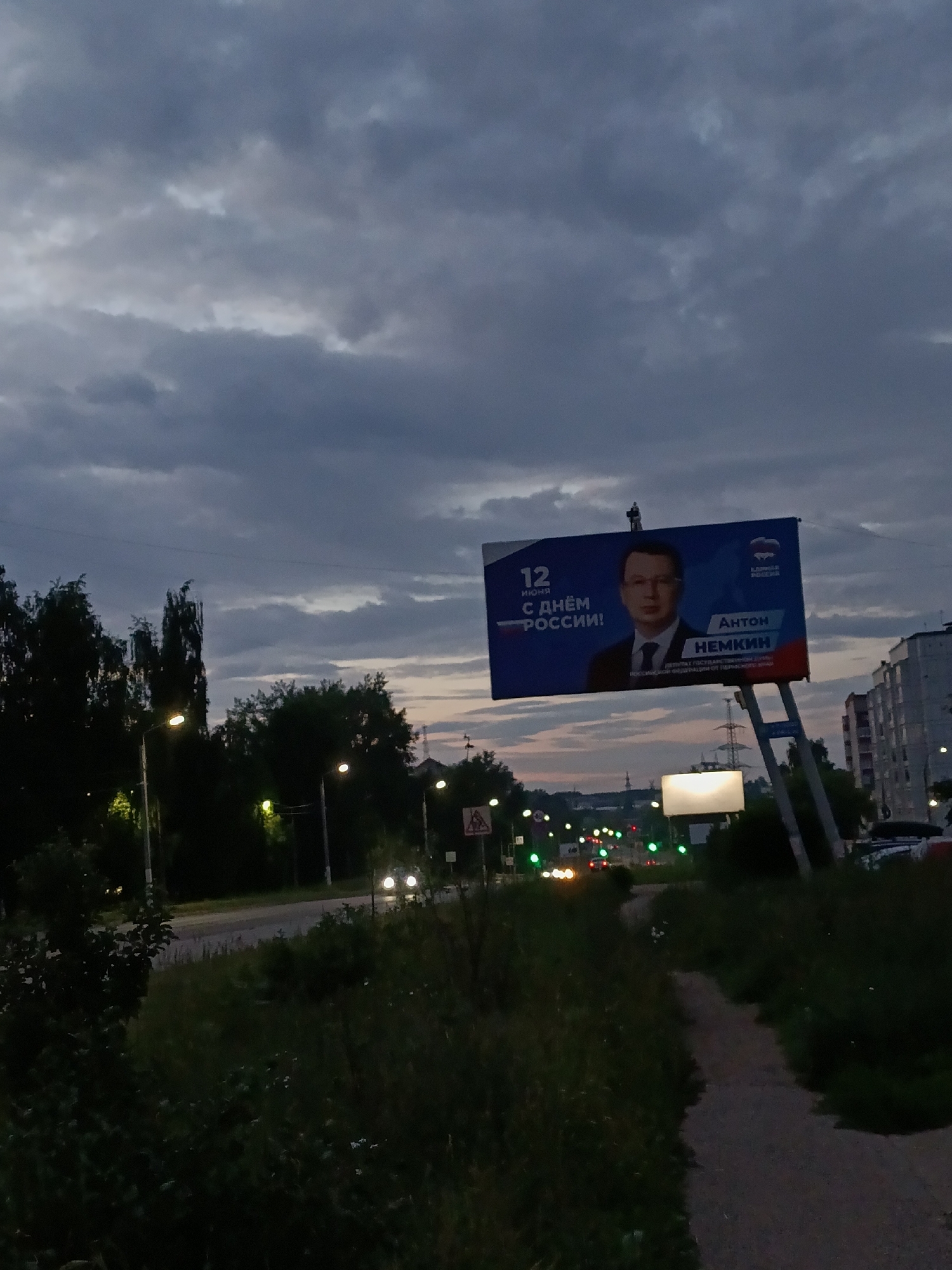 The image depicts a dusk scene in the city of Chaikovsky, Russia, featuring a large billboard by a road. The billboard displays a portrait of Anton Nemkin, identified as a deputy and member of the State Duma of the Russian Federation, with the text "12 ИЮНЯ С ДНЁМ РОССИИ!" (June 12, Happy Russia Day!). The sky is overcast with dramatic clouds, and streetlights illuminate the roadside. Trees line the left side of the road, and apartment buildings are visible in the background on the right. Traffic lights and distant vehicles suggest ongoing urban activity. The foreground shows a dirt path winding through overgrown grass.