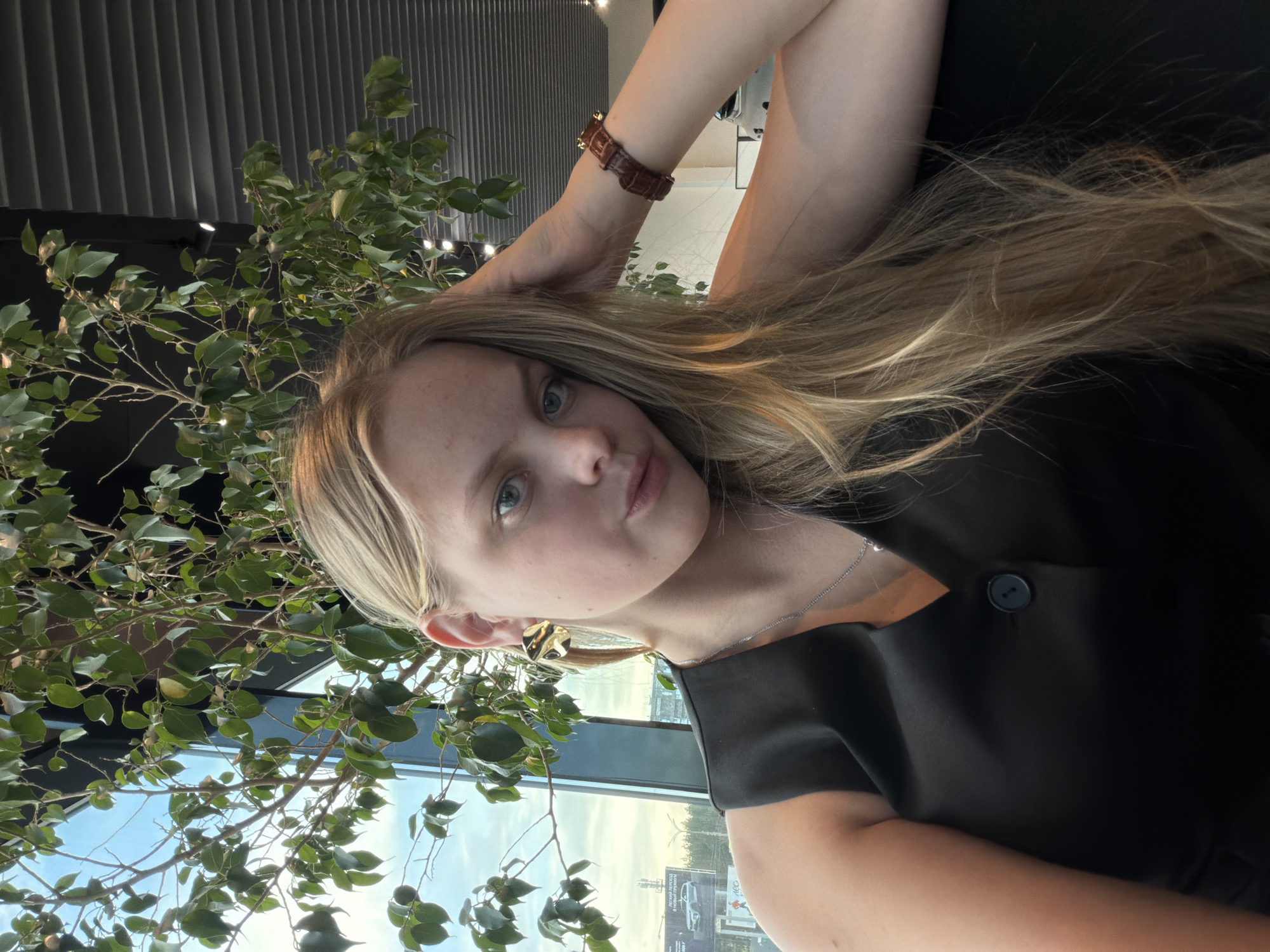 A woman with blonde hair is posing in front of a plant near a window. She is wearing a black top and a brown wristwatch. The background shows a cityscape that appears to be in Izhevsk, Russia.