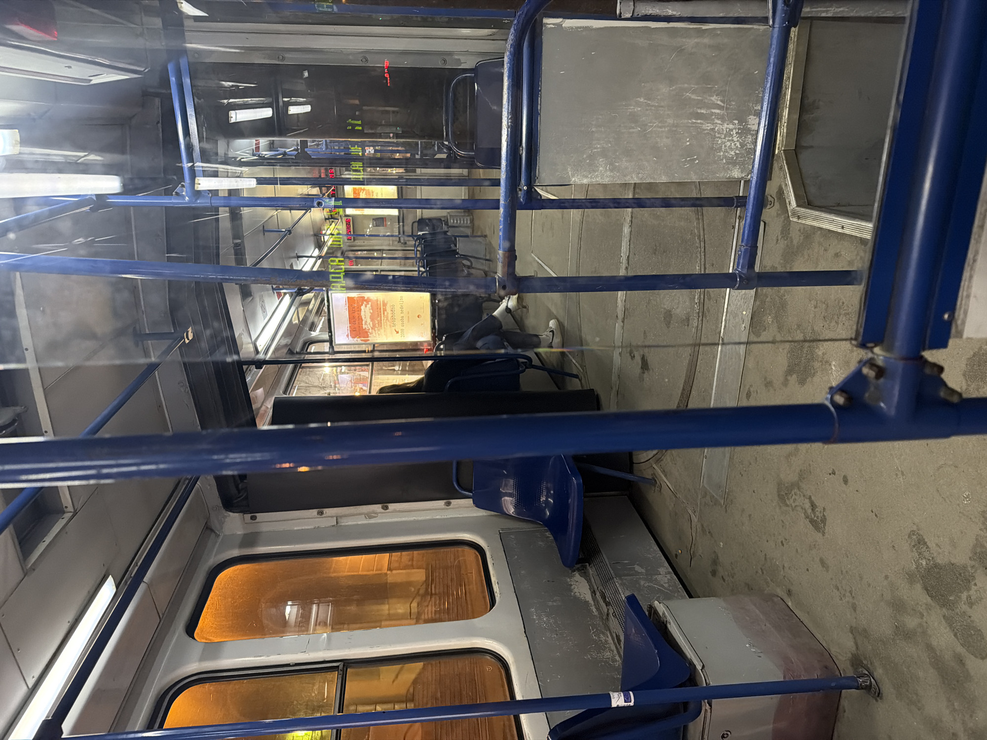 The image displays the interior of a public transport vehicle, specifically a tram, at night in Belgrade, Serbia. The perspective is from a lower angle, looking forward within the car, with a prominent blue metal pole and glass partition in the immediate foreground.
One individual is visible in the mid-ground, seated on the right side of the tram, facing away from the camera. This person is wearing dark clothing, dark trousers, and white athletic shoes (identifiable by a visible Nike swoosh on the left shoe) over light-colored socks. Their legs are casually extended.
The tram's interior features a utilitarian design. The floor is dark, appearing to be a worn, industrial-grade material with visible scuff marks and dirt. Numerous blue plastic bucket seats are arranged throughout the vehicle, with many appearing unoccupied. Blue metal poles and handrails, some showing wear, are distributed for passenger support. Large rectangular windows with rounded corners line the left side, reflecting interior lights and showing a blurry exterior environment characterized by warm orange and yellow streetlights, indicating nighttime. The walls and lower panels are light grey and exhibit signs of wear, including scuffs, scratches, and discoloration. Overhead, rectangular fluorescent light fixtures illuminate the interior from the light grey ceiling panels.
On the left side of the vehicle, through a glass barrier, a vertical poster with Serbian text is partially visible. Decipherable phrases include "ŠTO DAJE KREVETU" (What gives the bed), "neophodno" (necessary), and "2000 osoba nedeljno" (2000 people weekly), suggesting a public awareness or donation campaign. Reflections of interior lighting and elements are visible on the various glass and polished surfaces throughout the tram. A step-down section of the floor is visible towards the rear of the visible passenger compartment.