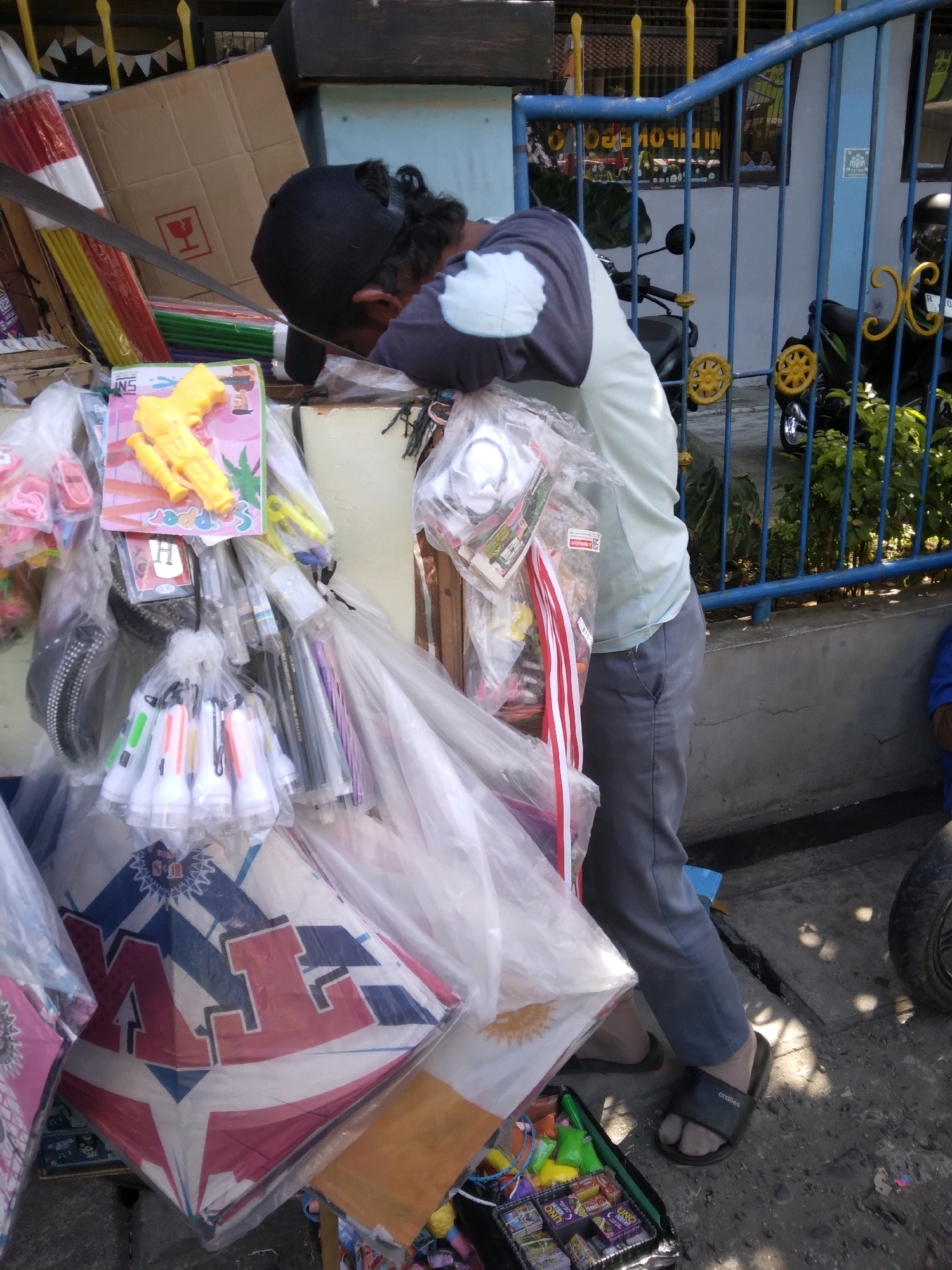 A person wearing a black baseball cap and a light blue and grey long-sleeved shirt is leaning over a display of goods. The person is wearing grey pants and black sandals. The display includes various items such as a yellow toy gun, packs of small flashlights, kites, and other packaged goods. The scene appears to be outdoors, with a blue metal fence and foliage visible in the background. A motorcycle is parked behind the fence. The ground is concrete with some cracks and debris. The overall impression is of a market stall or roadside vendor. The image was taken in Purwokerto, Indonesia.