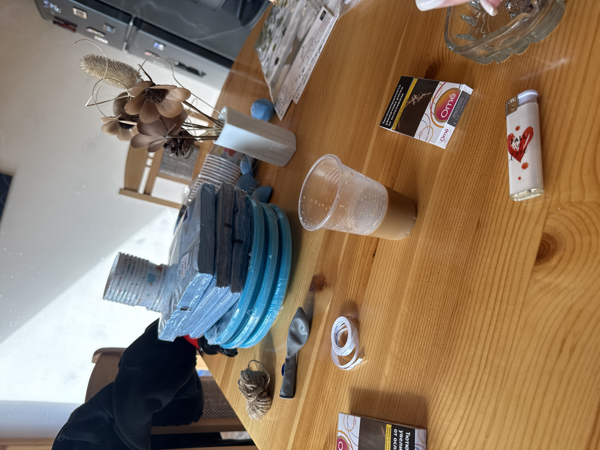 In Rogosh, Bulgaria, a close-up, high-angle shot captures a wooden table laden with various objects. In the foreground, a pack of "Omé" cigarettes rests beside a disposable plastic cup containing a light brown liquid, possibly coffee or iced tea. To the left, another "Omé" cigarette pack is partially visible. A white lighter with a red heart design and a few smoldering cigarette butts in a glass ashtray are situated to the right.
Further back, a deflated gray balloon lies near a spool of white tape. A ball of twine sits to the left of the balloon. A stack of blue disposable plates is prominently featured in the mid-ground, topped with a stack of white paper cups. A vase containing dried flowers and a pinecone stands behind the plates and cups. In the background, a glimpse of a chair and a refrigerator is visible, suggesting an indoor setting. The overall lighting suggests daylight, with shadows cast across the table.