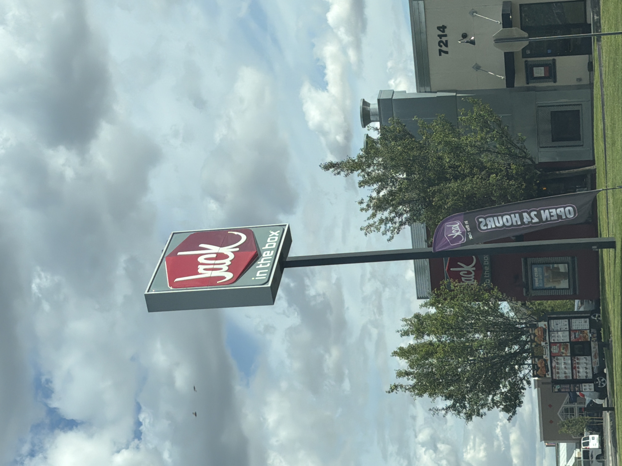 The image captures a Jack in the Box restaurant on a cloudy day in Pasco, Washington, United States of America. A prominent red and white Jack in the Box sign stands tall on a pole in front of the restaurant. To the left of the sign, a menu board displays various food items. To the right, a building with the number "7214" is visible, along with a dark awning and glass doors. A purple flag with the Jack in the Box logo is also present. The environment includes lush green trees and a paved area, likely a parking lot or drive-thru lane. The sky is filled with dramatic, scattered clouds.