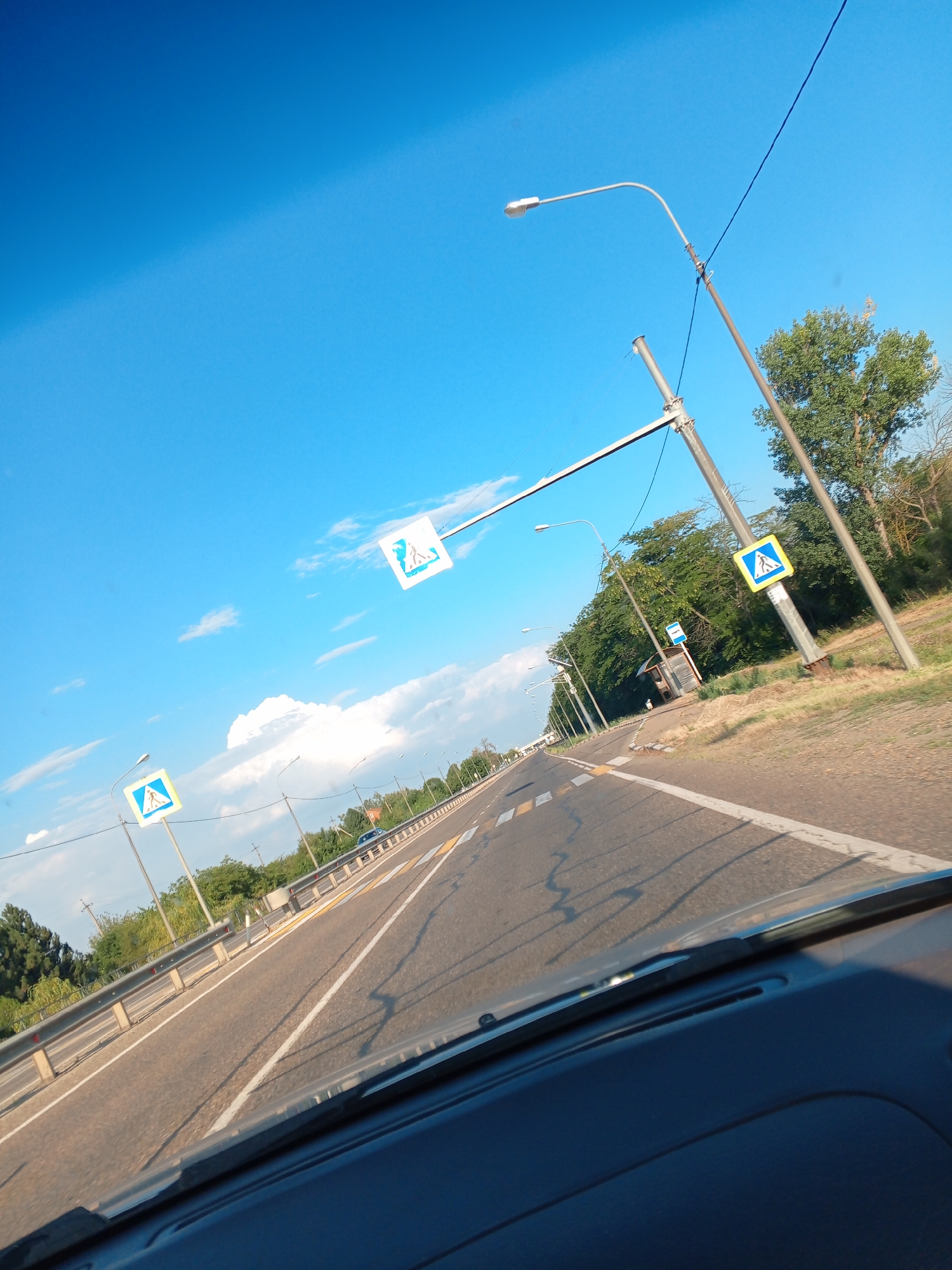 This is a first-person, dashboard-view photograph taken from a vehicle traveling on a road in or near Kamenny, Russia. The sky is a clear blue with scattered white clouds. A road stretches ahead, with a metal guardrail along the left side and trees lining the right side. Several pedestrian crossing signs, featuring a blue triangle with a white silhouette of a person walking, are visible along the roadside. There are also streetlights and utility poles. The road surface appears to be asphalt, marked with white lines. In the distance, a bus stop shelter is visible on the right. The interior of the vehicle, including the dashboard and windshield wipers, is partially visible in the foreground.