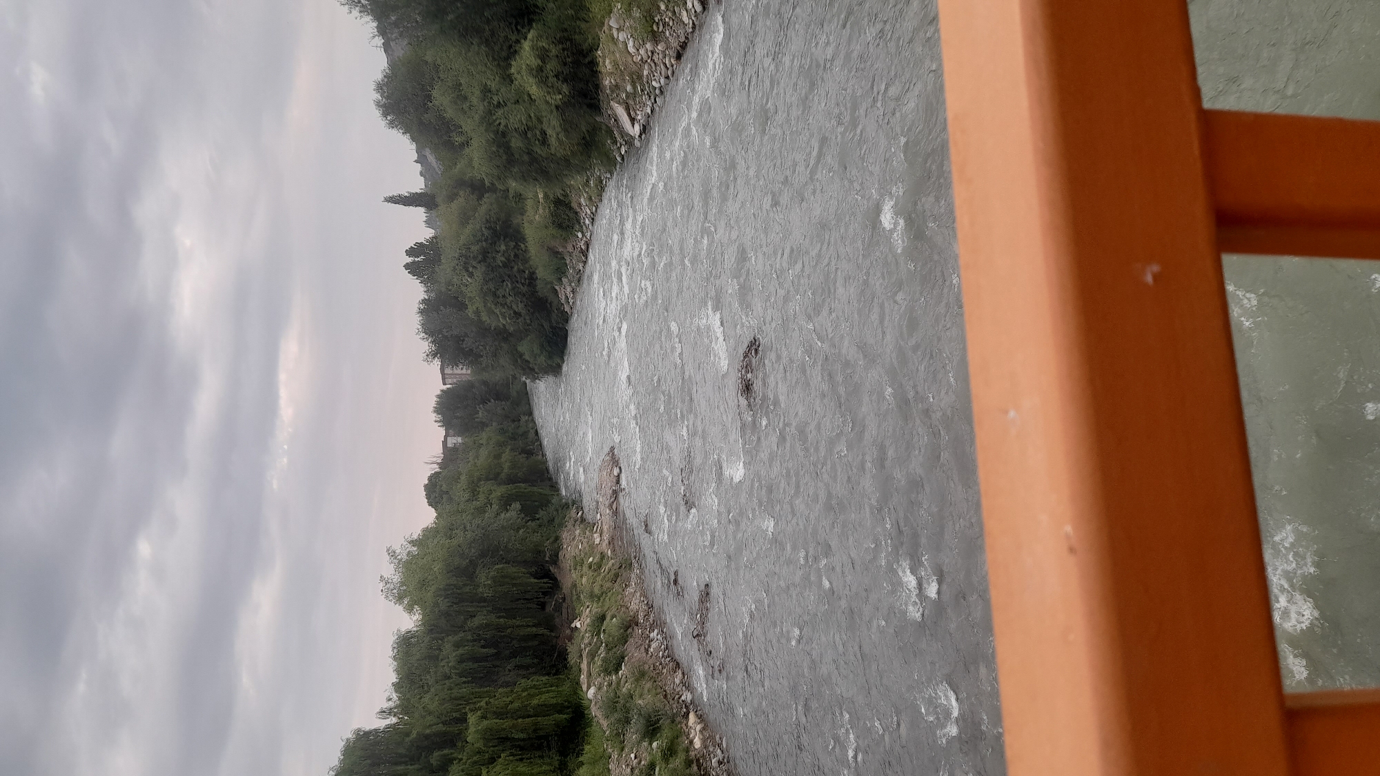 The image shows a view from a bridge railing overlooking a wide, fast-flowing river. The riverbanks are lined with dense green trees and vegetation. In the distance, there are some buildings visible among the trees. The sky is overcast with gray clouds. The railing in the foreground is orange.