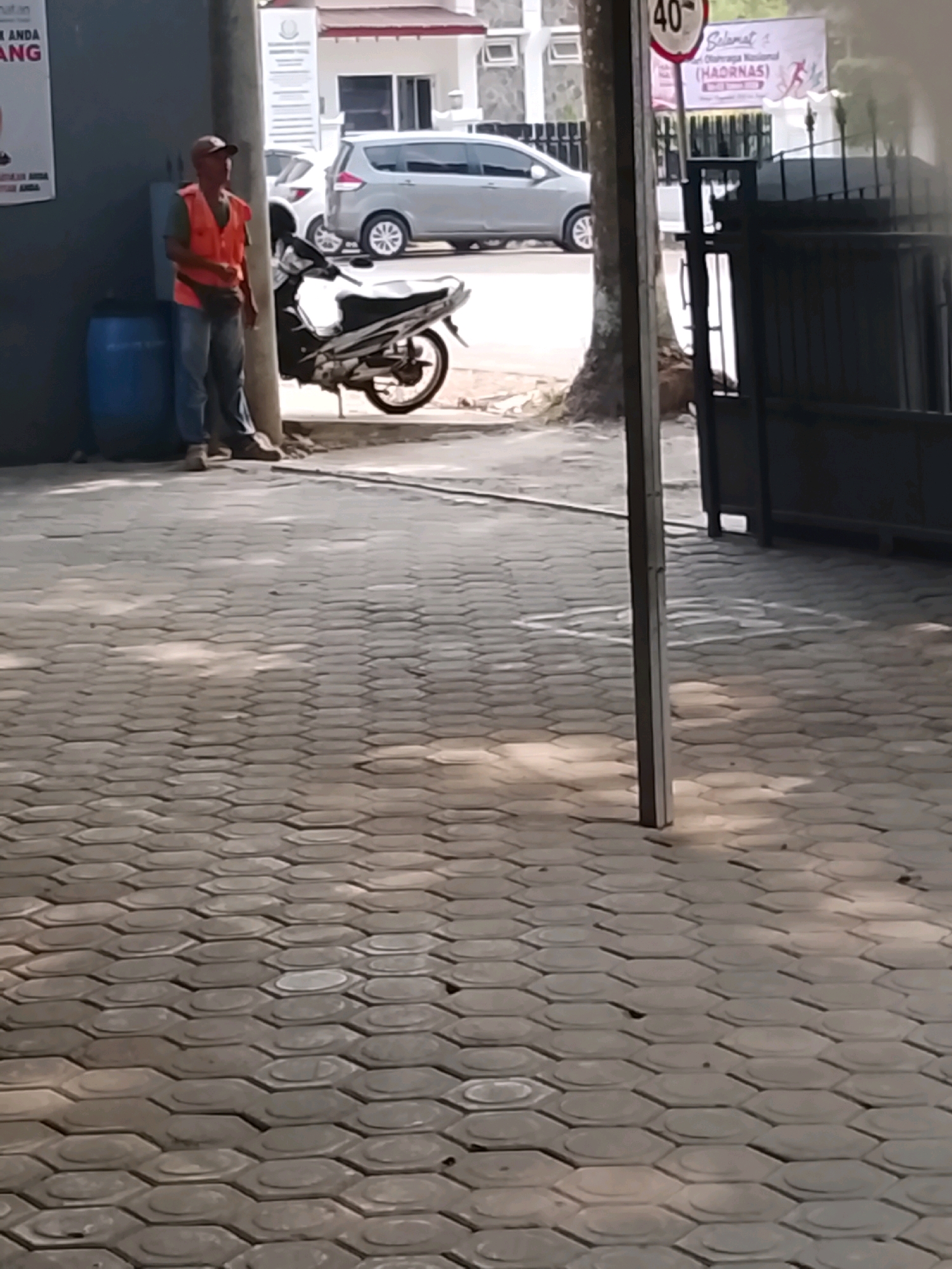 A man wearing an orange safety vest and a brown cap stands next to a blue barrel and a motorcycle. He is wearing blue jeans and brown shoes. The man is facing left and appears to be looking at something off-camera. Behind him, a silver car is parked on a street. A white building with a red roof is visible in the background. The image appears to have been taken in an urban setting in Slawi, Indonesia, as indicated by the context. The ground is paved with hexagonal tiles.
