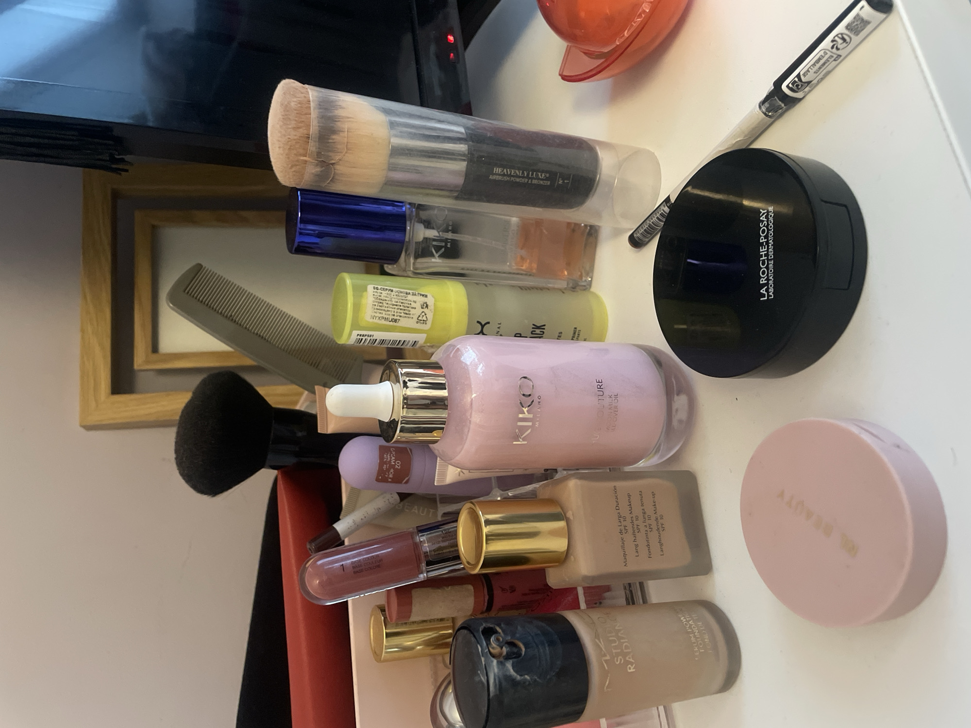 The image displays a close-up, top-down view of various cosmetics and beauty tools arranged on a light-colored surface, likely a dresser or table, in an indoor setting in Sofia, Bulgaria. The items are scattered but generally organized, suggesting a personal collection of makeup.
Key objects include:
* A large makeup brush in a clear plastic case, labeled "HEAVENLY LUXE® AIRBRUSH POWDER & BRONZER".
* Several foundation bottles, including a light beige one with text like "STUDIO RADIANCE SERUM POWER FOUNDATION" and indications of "Maquillaje de Larga Duración SPF 10", "Lang haftendes Makeup SPF 10", "Fondotinta a lunga tenuta SPF 10", and "Langhoudende Make-up SPF 10".
* A light pink bottle with a dropper, branded "KIKO MILANO PURE COUTURE OIL". Another smaller purple tube is visible with "BASE COULEUR" and "02".
* A black compact powder case, clearly marked "LA ROCHE-POSAY LABORATOIRE DERMATOLOGIQUE".
* A round, pale pink compact powder case, labeled "RARE BEAUTY".
* A yellow bottle with "PREP SET" and partially visible text "ВО-СЕРУМ ОСНОВА ЗА ГРИМ", which is Bulgarian for "BO-SERUM MAKEUP BASE", confirming the context of Sofia, Bulgaria. It also has "NYXPMU087".
* Several lip products, including a clear lip gloss with "BASE COULEUR" visible.
* A black hair comb and a smaller black makeup brush with a rounded head.
* A black pen or pencil with "ÉLÉMENTS D'EMBALLAGE" (packaging elements) and a recycling symbol.
In the background, part of a television or monitor and a wooden picture frame can be seen. No specific activity is depicted other than the arrangement of these personal care items.