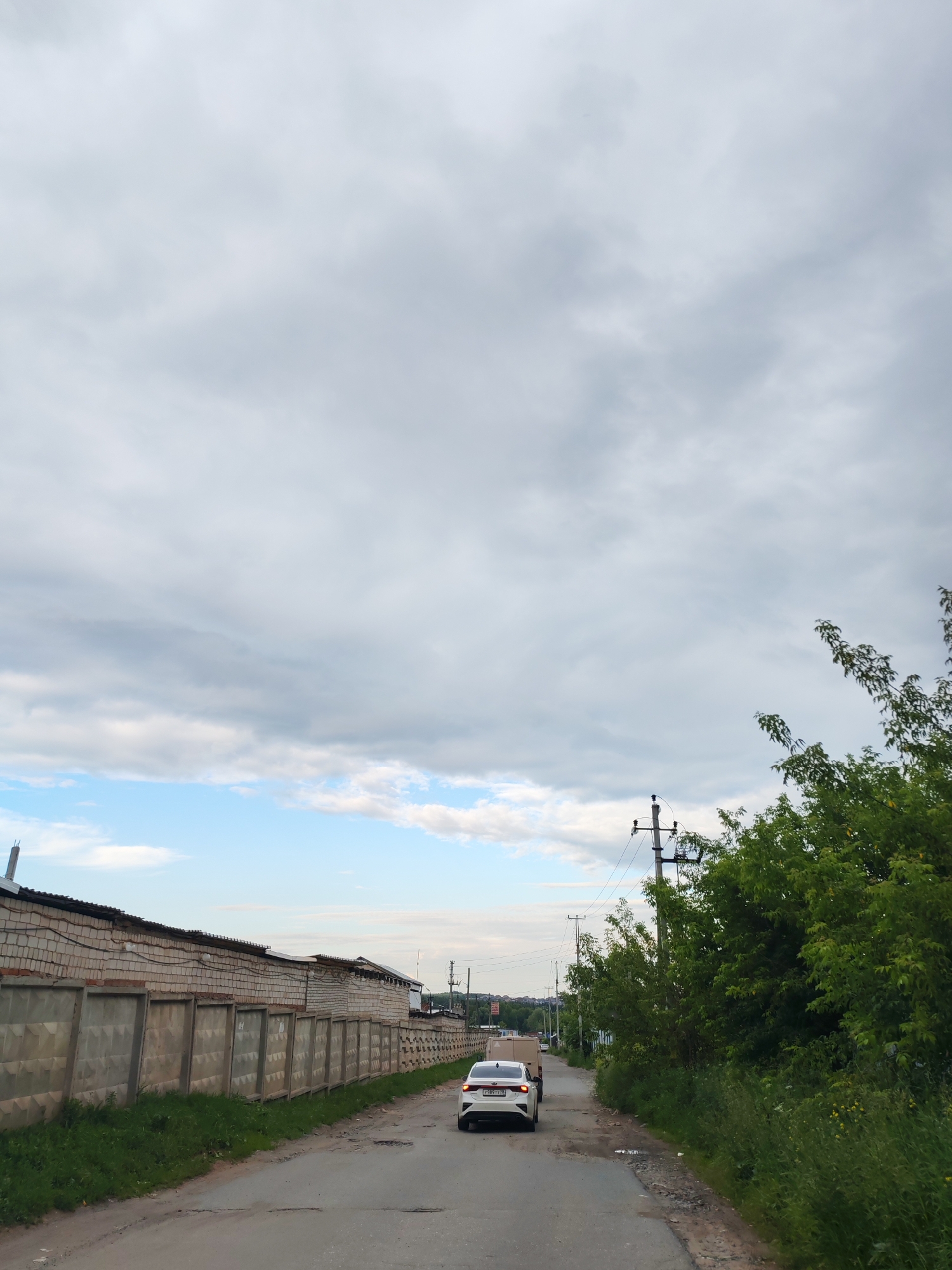 The image shows a grey, cloudy sky over a road in Izhevsk, Russia. A white car is driving down the road, and there is a long concrete fence on the left side of the road. Trees and greenery are visible on the right side of the road. Power lines and poles are also present. The scene appears to be a rural or industrial area on the outskirts of the city.