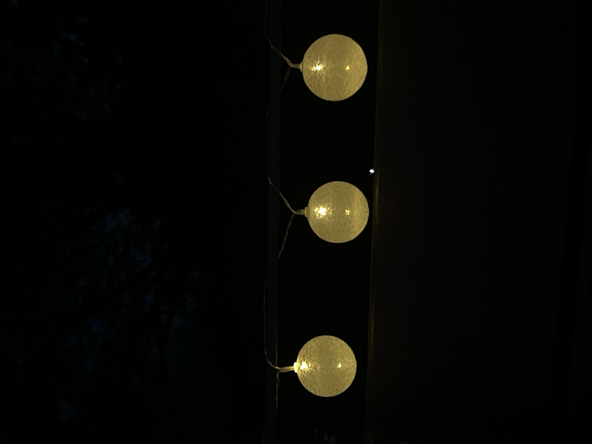 Three round, yellow string lights are illuminated against a dark background. The lights are suspended from a thin wire that stretches horizontally across the frame. Each light appears to be made of a woven material and contains a single bulb that emits a warm glow. The background is uniformly dark, suggesting nighttime. The location is not discernible from the image.