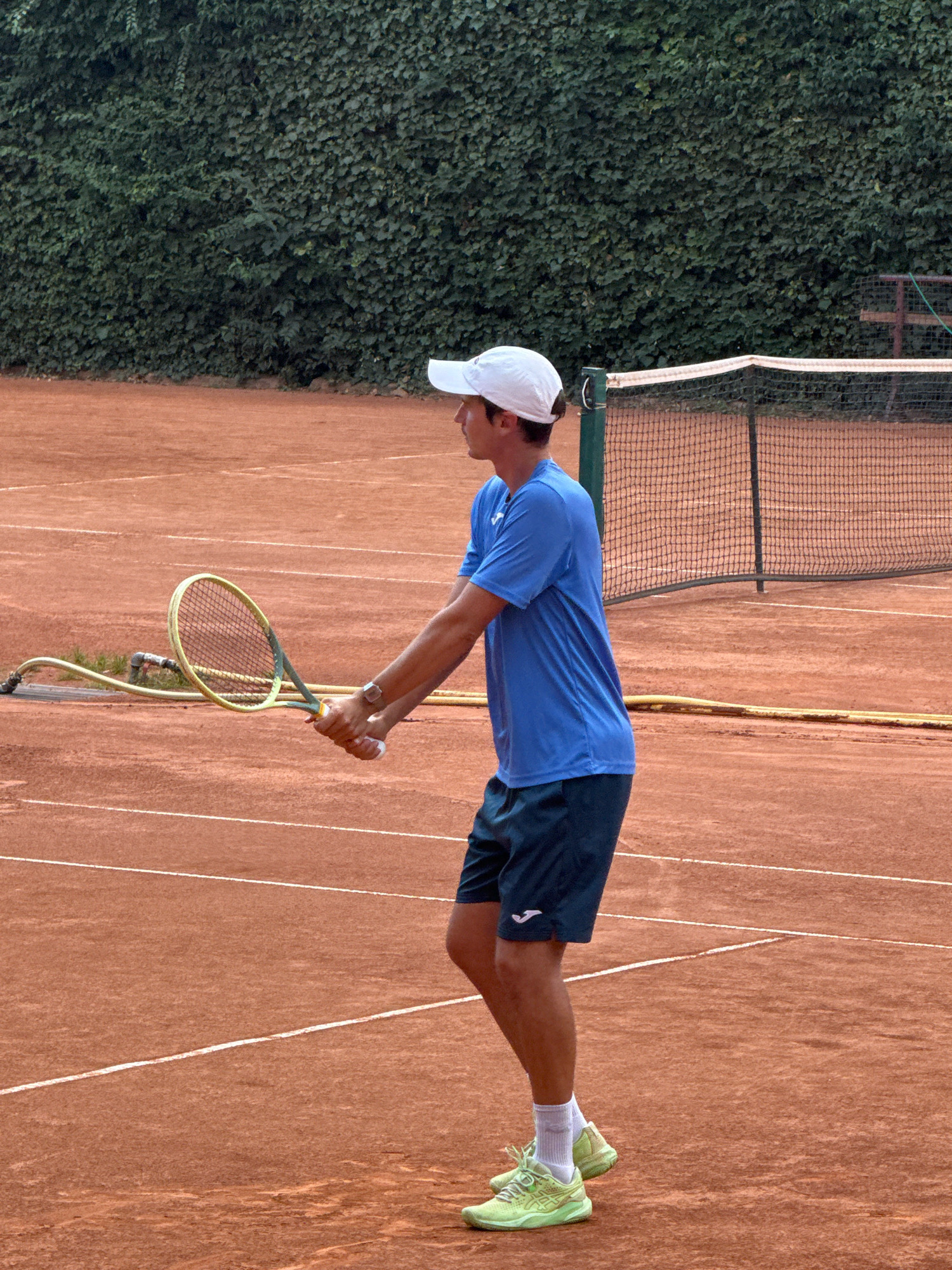 A young man, likely in his late teens or early twenties, is positioned on a clay tennis court, mid-action in preparation for a shot. He is wearing a blue short-sleeved t-shirt, dark blue shorts, a white baseball cap, white socks, and light green tennis shoes. He holds a tennis racket in both hands, with the racket face tilted downwards. A watch is visible on his left wrist. The court is a reddish-brown clay surface with white boundary lines. In the background, a tennis net is strung across the court, and beyond that, a dense green hedge serves as a backdrop. A yellow hose lies coiled on the court near the net post on the left side of the frame. The lighting suggests an outdoor daytime setting, possibly overcast or in shade. The overall scene depicts a moment of athletic concentration during a tennis match or practice.