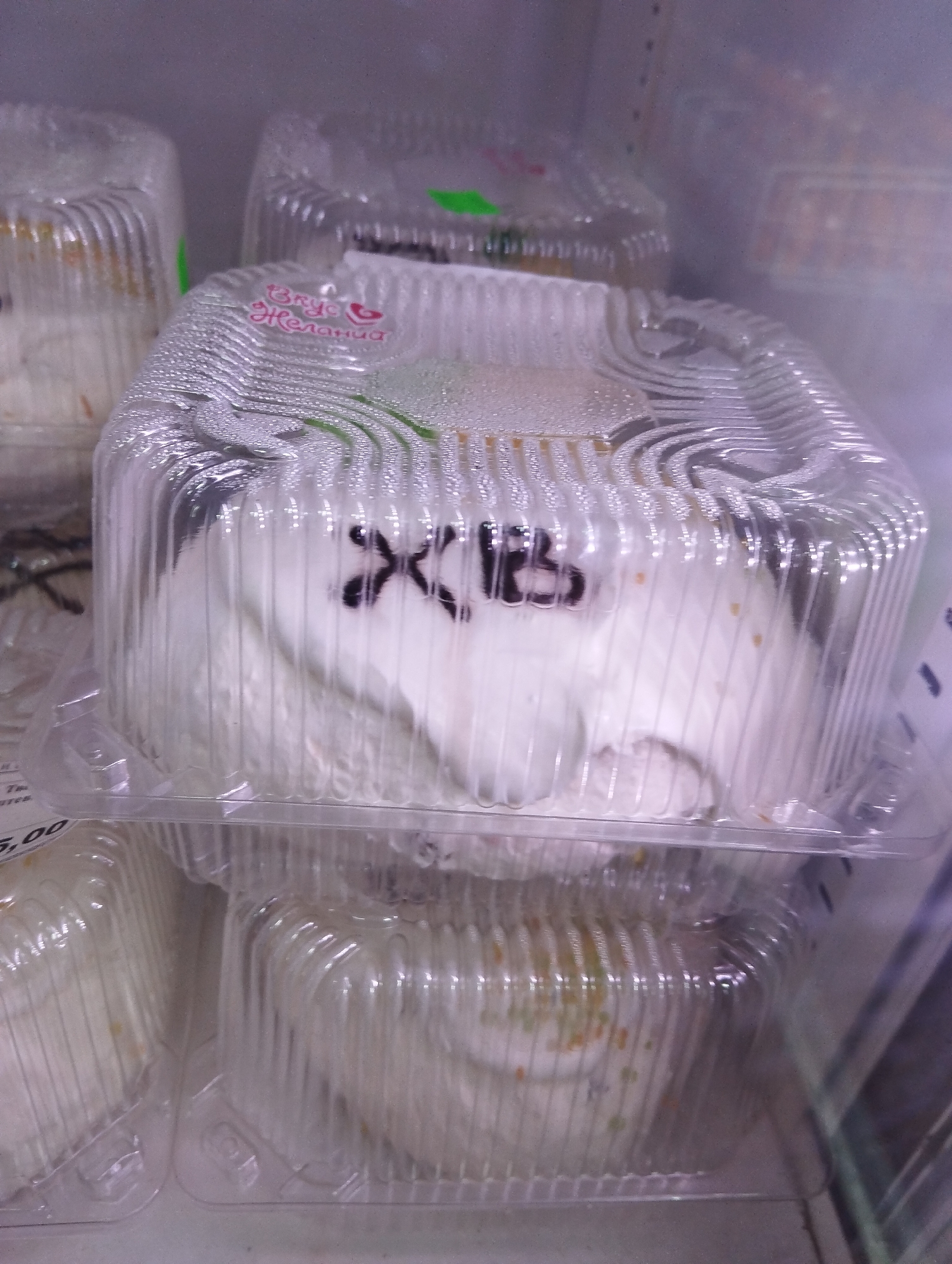 In a refrigerated display, presumably in a shop in Krasnogorskoye, Russia, there are several square, transparent plastic containers stacked on shelves. Each container holds what appears to be an Easter cake, covered in white icing with the Cyrillic letters "XB" written on top in dark icing. The top container is in focus. The cakes are frosted and are likely for sale. Some containers have condensation on their surfaces. A pink logo of “Вкус Желания” (Taste of Desire) can be seen on the packaging. A price sticker is visible on the shelf below. The environment seems to be a shop setting.