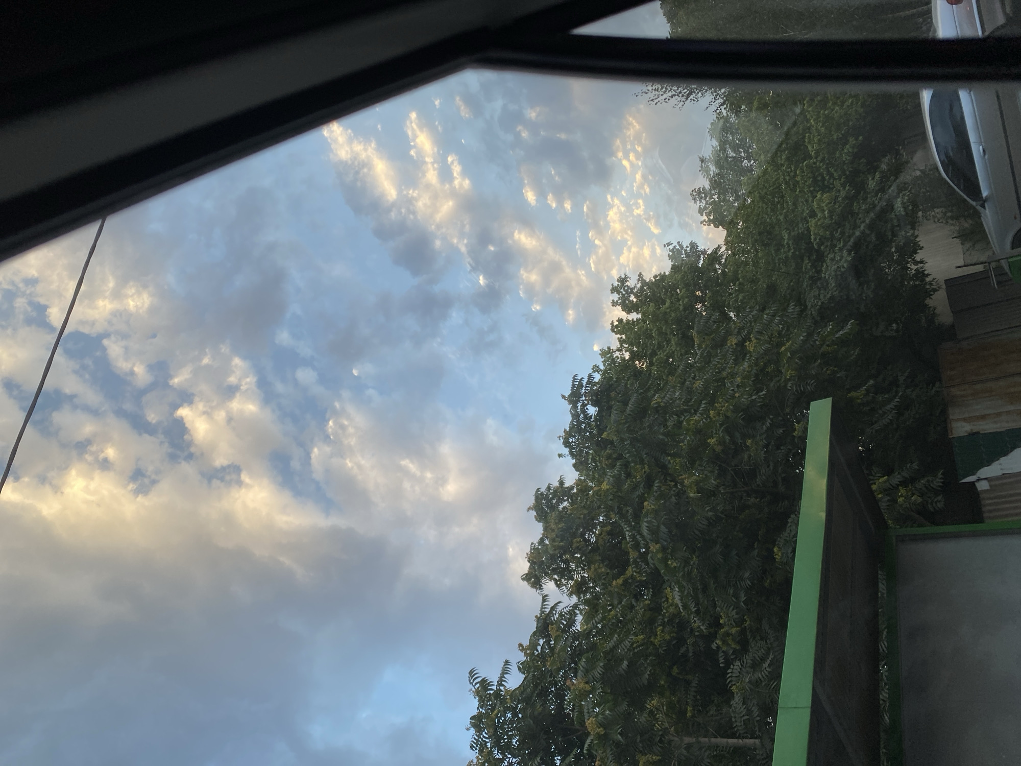This is an eye-level, medium shot photo taken from inside a vehicle looking out at a bus stop shelter and trees under a cloudy sky. The sky is a mix of blue and white clouds, with some areas illuminated by the sun, suggesting it is either early morning or late afternoon. A green bus stop shelter with a metal roof is visible on the left side of the frame. Behind the shelter, dense green trees with dark leaves fill the middle ground. In the background, a silver car is parked on the right side of the frame. The scene appears to be in an urban or suburban environment, likely Bishkek City, Kyrgyzstan, given the provided context, depicting an ordinary street view.