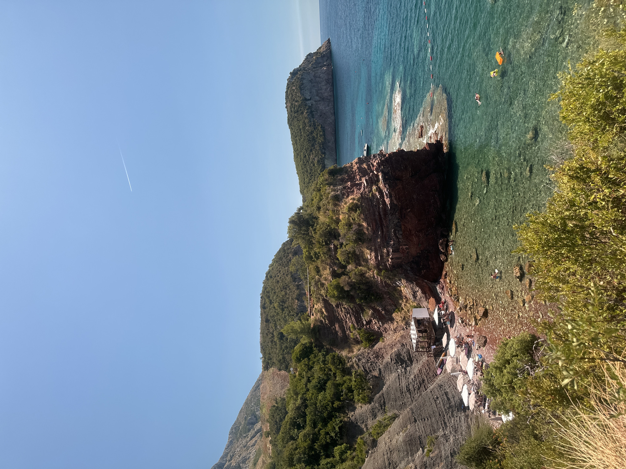 The image depicts a scenic coastal view in Sveti Stefan, Montenegro, featuring a rocky shoreline with a small beach area. The beach is dotted with numerous white umbrellas and lounge chairs, suggesting a populated recreational spot. People are visible relaxing on the beach and swimming in the clear turquoise waters. Several boats are anchored offshore, and a small structure, possibly a beach bar or cafe, is situated near the edge of the water. The background is dominated by lush green hills and steep rocky cliffs that lead up to a distant mountain range. The sky is a clear, bright blue with a faint contrail from an aircraft. The water is calm and transparent, allowing visibility of the seabed with submerged rocks and pebbles. The scene conveys a tranquil summer atmosphere.