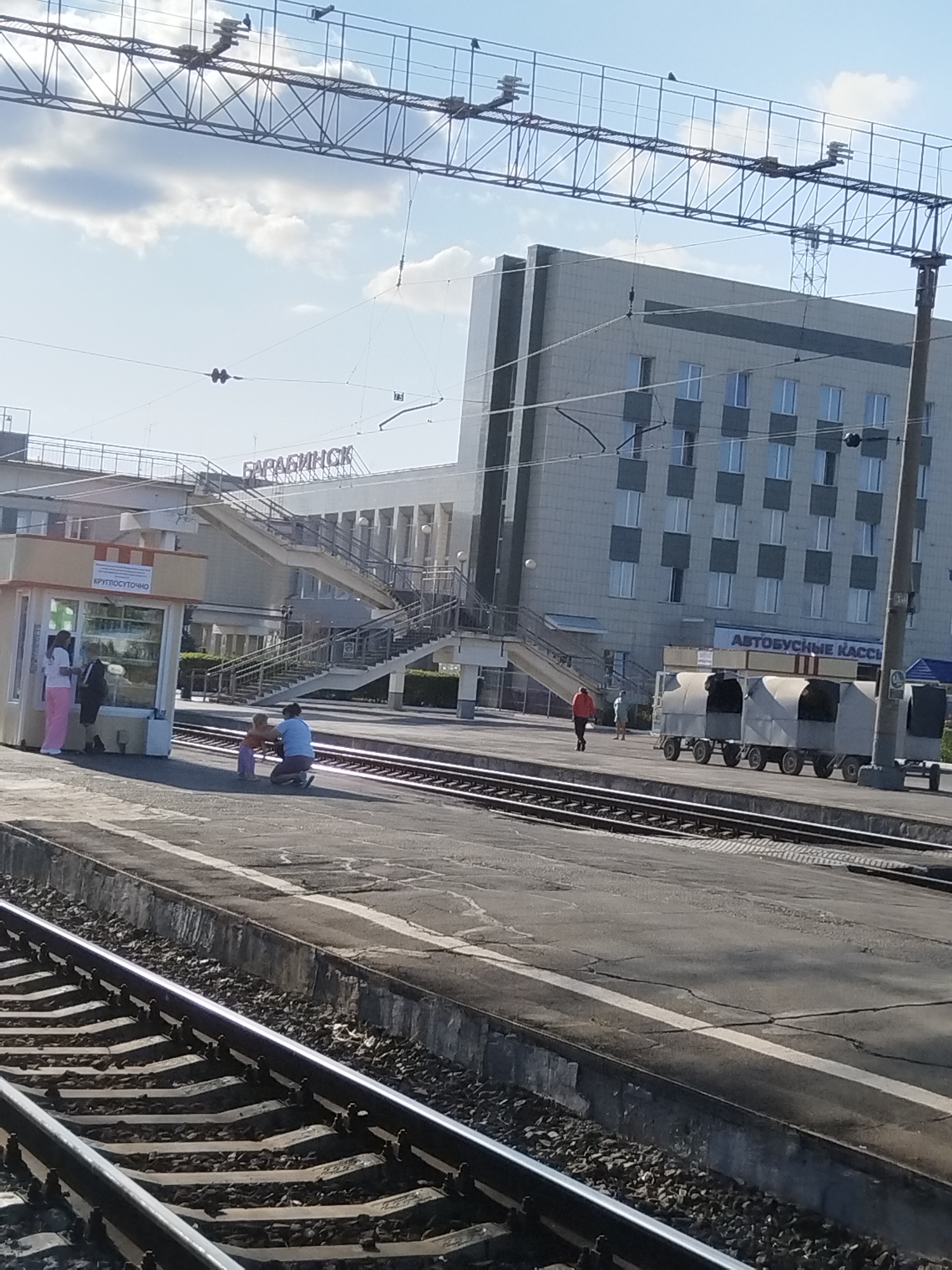 The image shows a train station in Barabinsk, Russia. In the foreground, there are train tracks. On the platform, a woman in a blue shirt and beige pants is kneeling and facing a child in an orange shirt. To the left of them, another woman in a pink shirt and pink pants is standing near a small kiosk that sells drinks. In the background, there is a large, modern building with multiple windows. Above the building, there are power lines and a sky with clouds. To the right of the building, there are several train cars parked on a separate track. A sign on the building reads "Автобусные кассы" (Bus ticket office). Another sign on the kiosk reads "КРУГЛОСУТОЧНО" (Around the clock).
