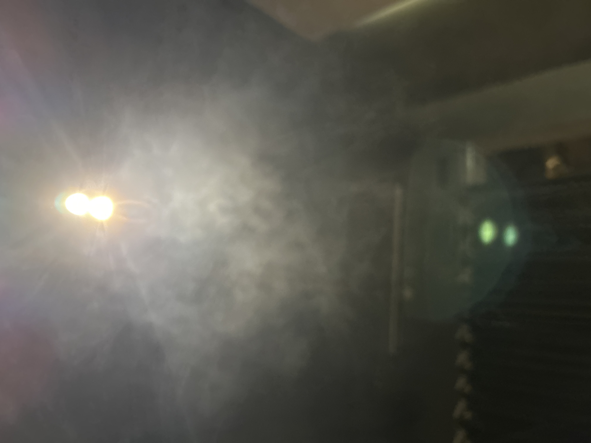 The image shows a hazy or smoky indoor environment. A bright light source is visible on the left side of the image, surrounded by a cloud of what appears to be smoke or fog. On the right side, there's a blurred, dark object with a reflective surface that shows two points of light. The background is mostly dark and indistinct. The location and specific details about people, subjects, objects, actions, and interactions are not discernible from the image.