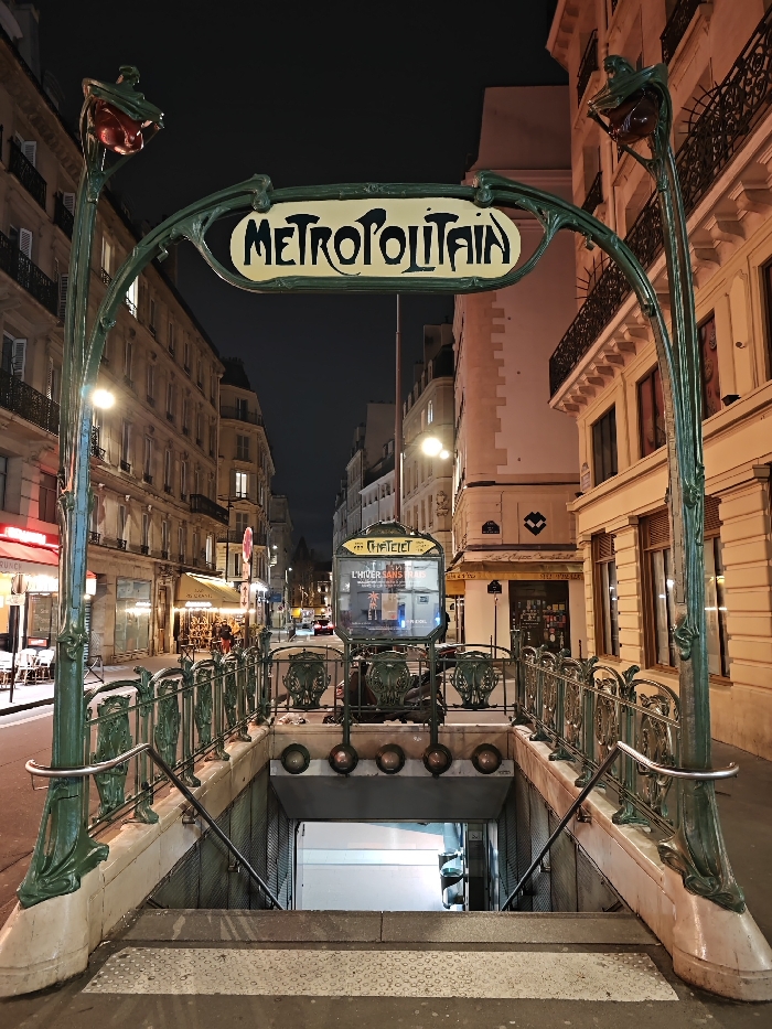 A Métropolitain entrance in Paris is captured at night. The iconic Art Nouveau sign is prominent. A wrought iron railing and light fixtures adorn the entrance. Steps lead down into the subway. Buildings line the narrow street, illuminated by streetlights. A Châtelet metro sign is seen behind the structure. The scene evokes a classic Parisian atmosphere.