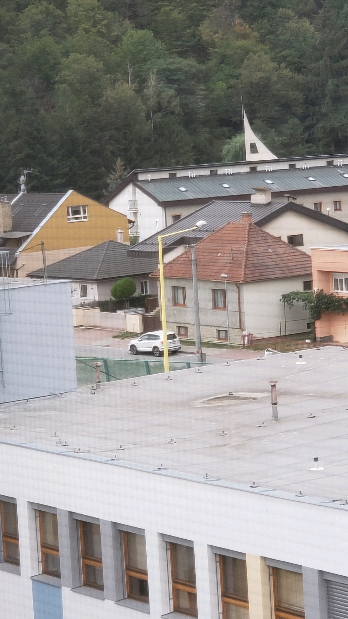 This image depicts a suburban residential area in Prešov, Slovakia, with a focus on a street scene and surrounding buildings. In the foreground, a flat, grey rooftop with visible metal fasteners and a few protrusions is prominent. Behind this rooftop, a white building with multiple rectangular windows is visible. Further back, a street curves, with a white SUV parked on its right side. Adjacent to the street is a red-tiled roof house with several windows and a chimney. A tall, yellow lamppost stands near the house. Several other houses and buildings with various roof types and colors are visible in the middle ground. The background features a dense forest covering a hillside. The sky is overcast, suggesting cloudy weather.