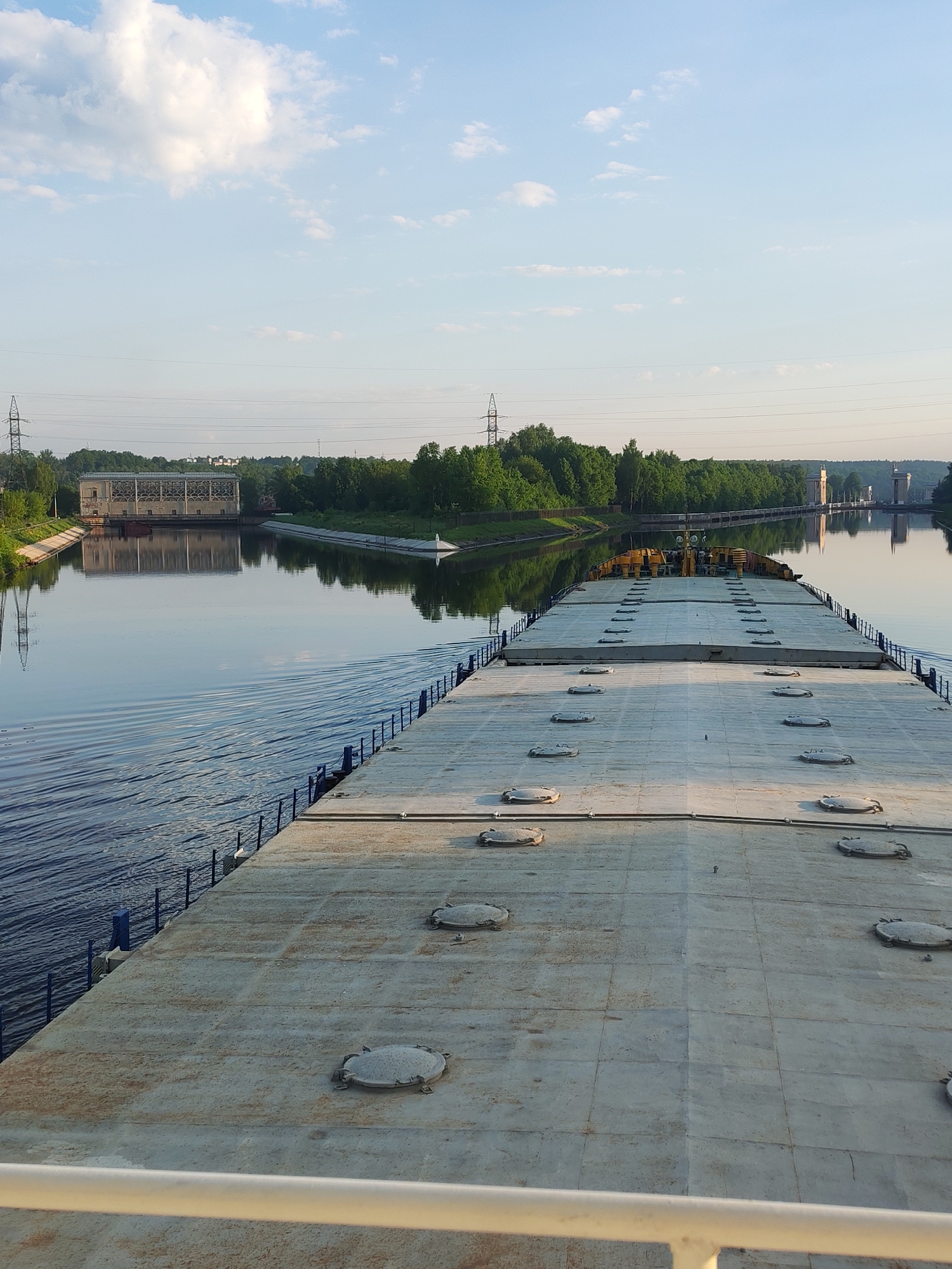 Here's a detailed description of the image:
The image appears to be taken from the deck of a large barge or ship traveling along a waterway near посёлок опытного хозяйства «Ермолино», Russia. The deck is gray, composed of rectangular sections with multiple circular access hatches. Blue railings run along the edges of the barge.
The waterway is wide, reflecting the sky. On the left bank, there's a large building, possibly a pumping station or industrial facility. A line of power transmission towers and lines run parallel to the waterway. Trees line both banks. A lock system is visible in the distance on the right.
The sky is blue with scattered white clouds, indicating a clear day. The lighting suggests it is either early morning or late afternoon.