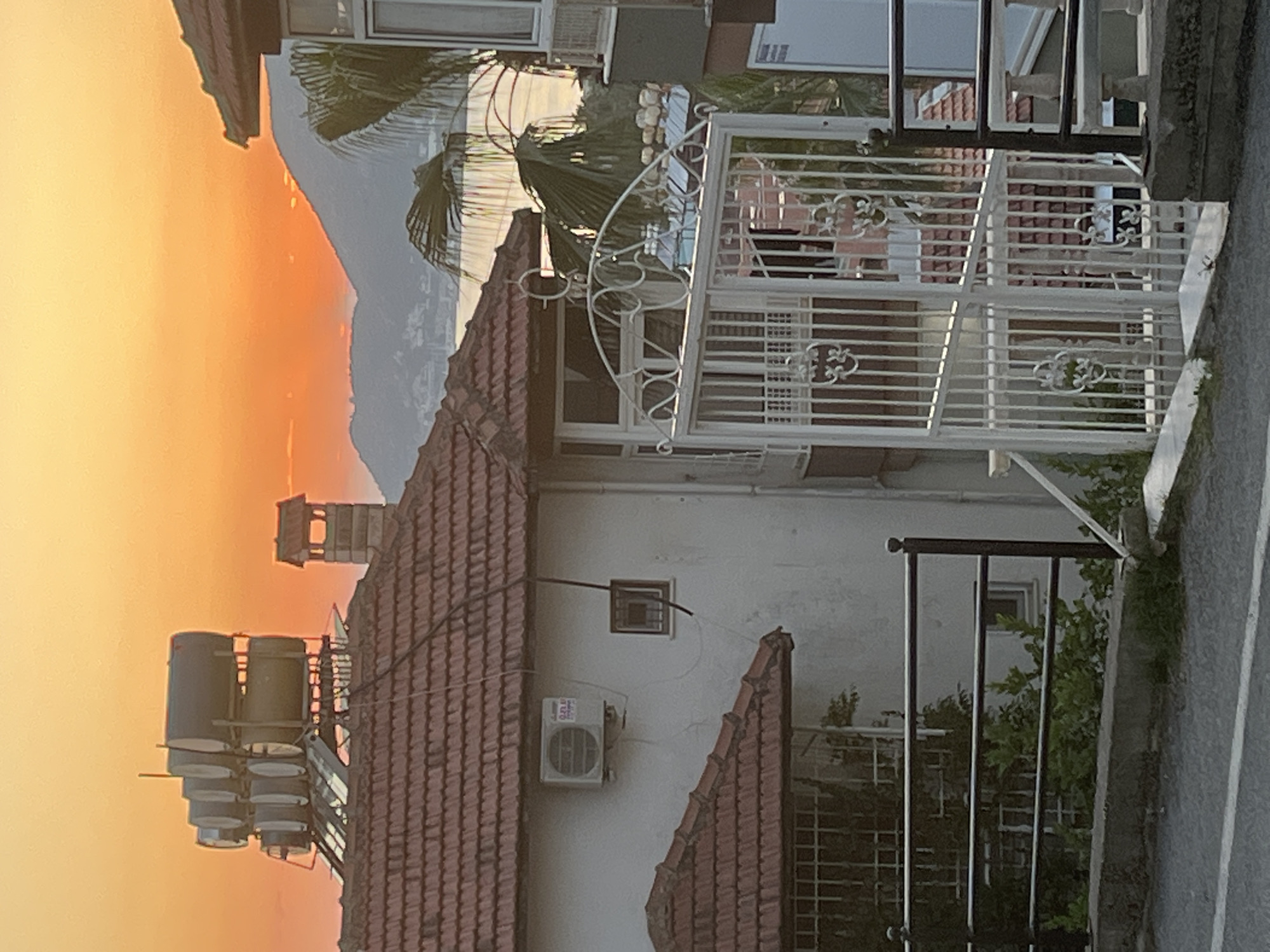 The image depicts a scene in Alanya, Turkey, during sunset. The sky is a gradient of orange and yellow. In the foreground, there is a house with a red-tiled roof. Attached to the roof is a solar water heating system consisting of multiple tanks. A white air conditioning unit is visible on the wall of the house. A decorative white metal gate is in front of the house with another fence beyond. The landscape includes palm trees. The background features mountains silhouetted against the sunset sky. A road is seen on the right.