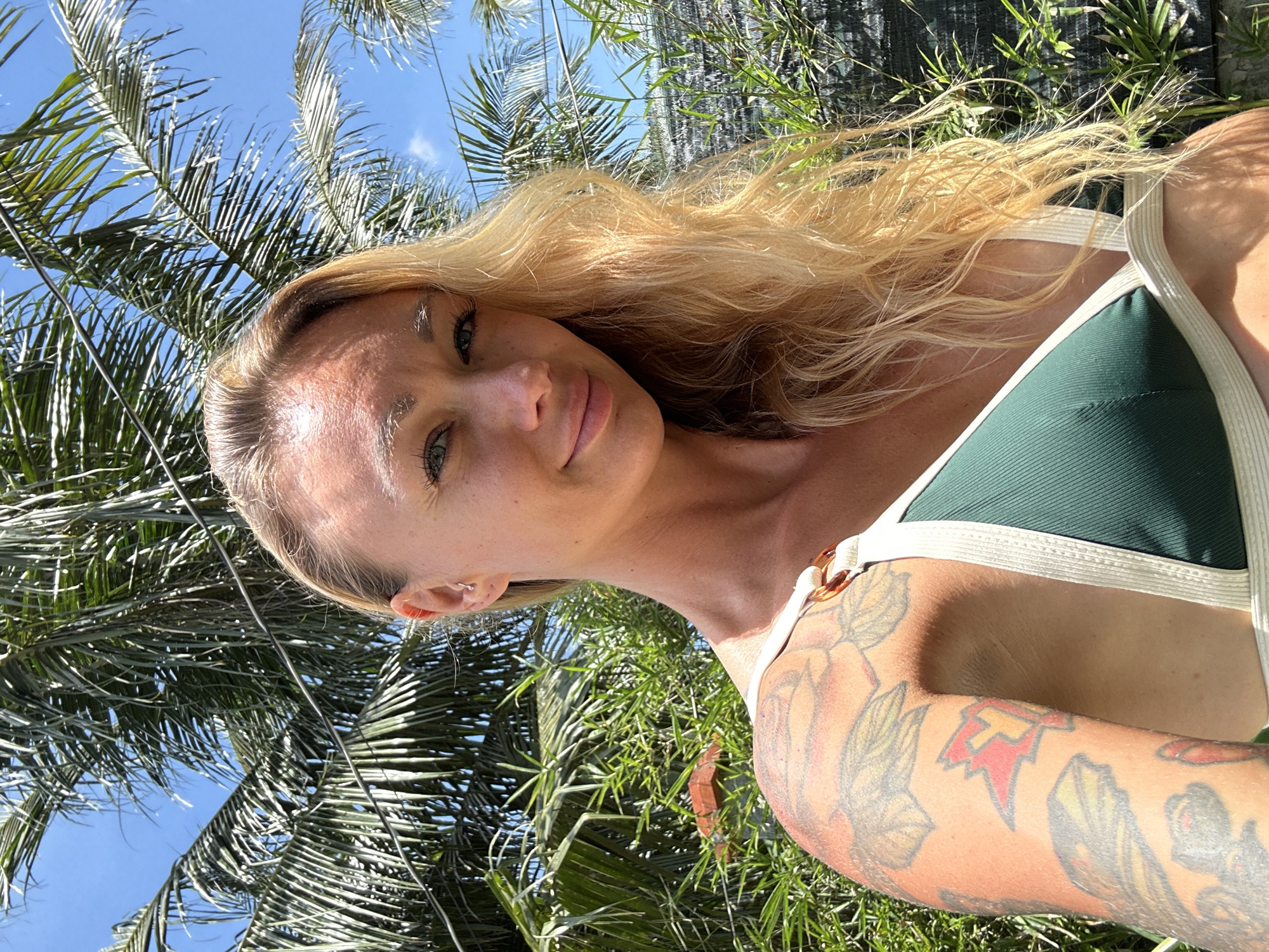 In Mũi Né, Vietnam, a woman with blonde hair and light green eyes poses for a selfie. She is wearing a dark green bikini with beige trim. She has tattoos on her left arm and shoulder. Behind her, palm trees and lush green foliage are visible against a bright blue sky. The scene suggests a sunny outdoor setting, possibly a resort or a beach area.