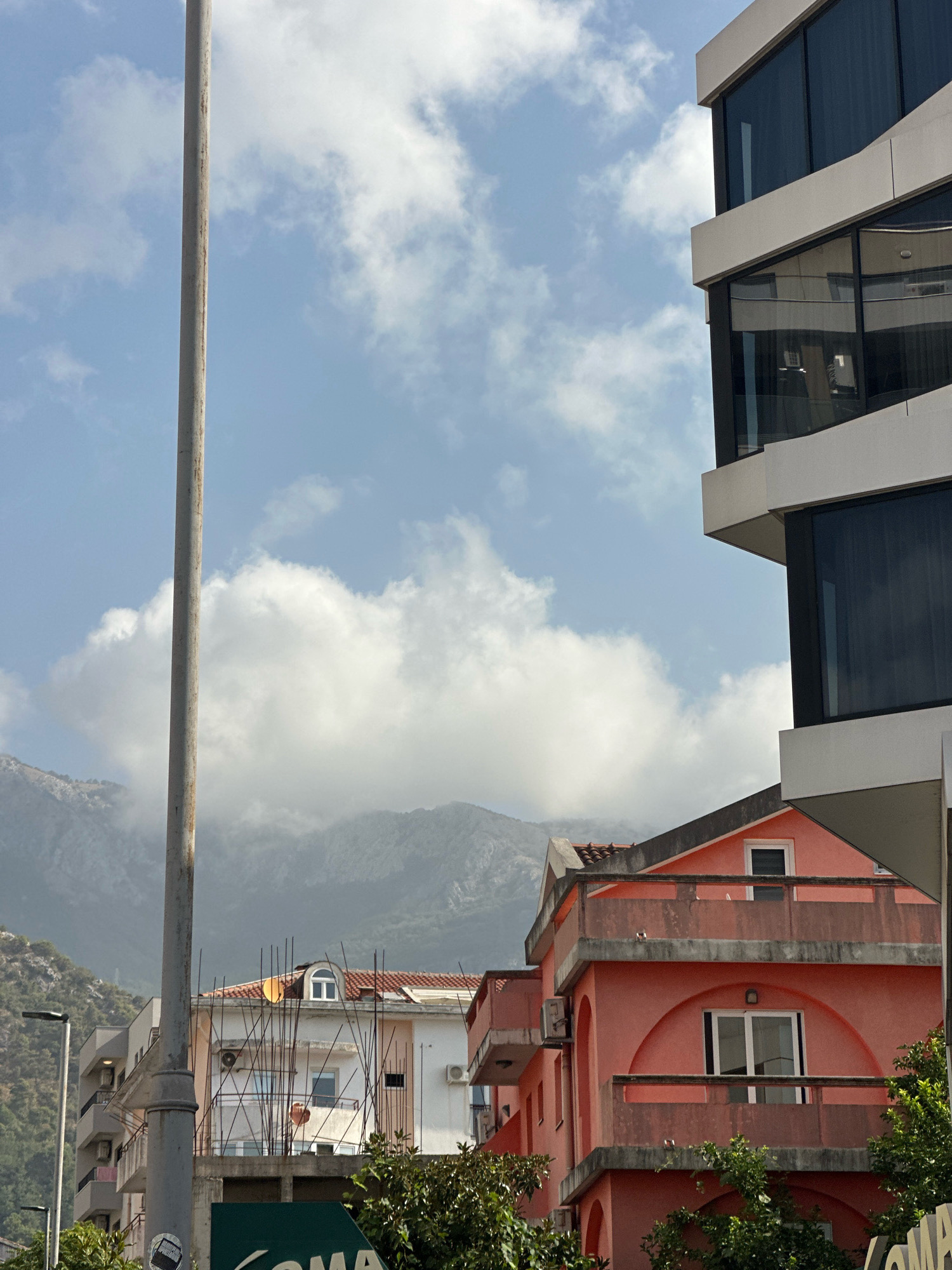 The image depicts an urban scene in Budva, Montenegro, with a clear sky and scattered clouds. In the foreground, a tall, grey utility pole stands prominently on the left side of the frame. To the right of the pole, a modern building with a white facade and dark, reflective windows juts into the upper right corner of the image. Below this, a brightly colored, pink residential building with balconies and arched window frames is visible. In the background, a white building with exposed rebar and satellite dishes suggests ongoing construction or renovation. Beyond the buildings, a range of mountains is shrouded in mist and low-lying clouds, adding depth to the landscape. The overall impression is one of a developing coastal town under a partly cloudy sky.