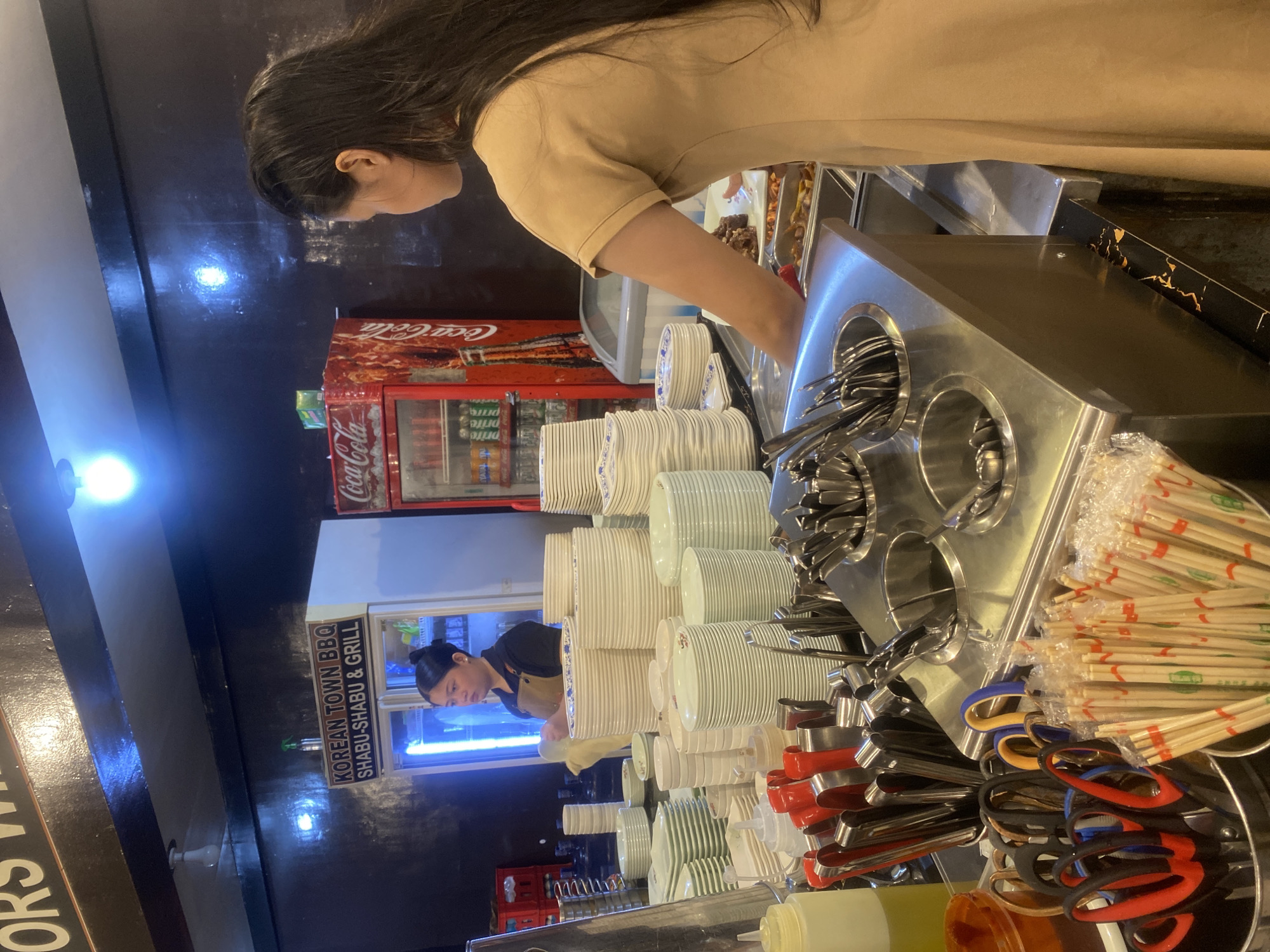 The image depicts an indoor scene at "KOREAN TOWN BBQ SHABU-SHABU & GRILL" in Davao City, Philippines. A woman with long, dark hair, wearing a tan-colored dress, is standing behind a stainless steel counter. She is holding a plate of food and appears to be serving or preparing it. In the background, a red Coca-Cola refrigerator is visible, stocked with beverages. Various stacks of white plates, serving utensils, and chopsticks are arranged on the counter. A woman in a black shirt and apron is standing near a refrigerator, also appearing to be an employee. The environment suggests a restaurant setting, likely a buffet or self-service establishment.