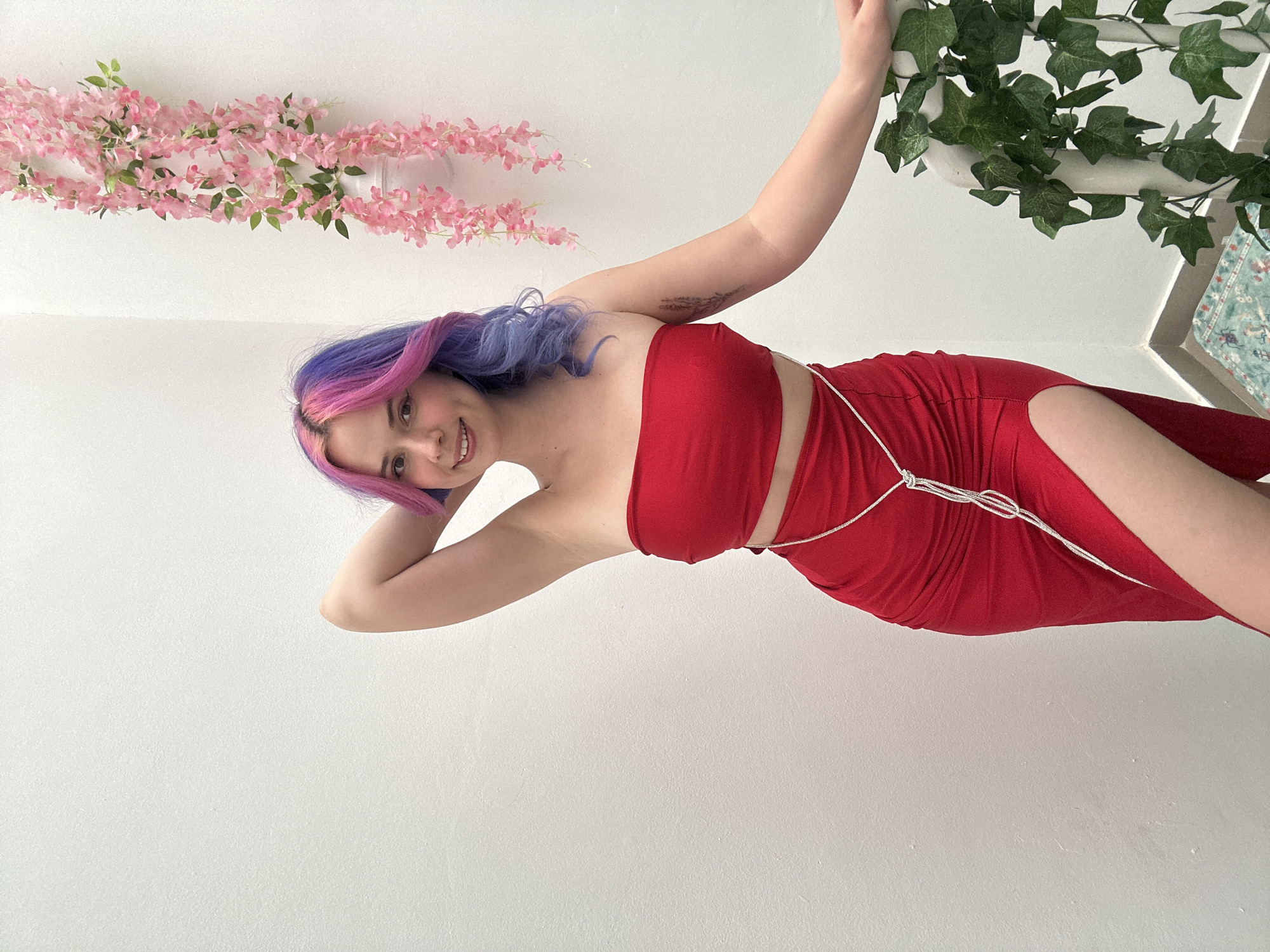 A young woman is prominently featured in an indoor setting, with the image rotated 90 degrees clockwise. She has striking bi-colored hair, transitioning from vibrant pink at the roots to purple along the length. She is wearing a strapless, form-fitting red dress that includes a high slit on the right side and is adorned with a delicate, silver-colored, possibly rhinestone, string detail wrapped around her waist.
The woman is smiling broadly at the viewer. Her left arm is extended upwards (to the right in the rotated image), appearing to hold onto a cluster of artificial green ivy leaves that are attached to a white surface. Her right arm is bent, with her hand near her head. A faint tattoo is visible on her left upper arm.
The background consists primarily of plain white walls and ceiling. In the upper left (bottom left in the rotated view), a garland of artificial pink flowers with small green leaves is strung across the white surface. A small portion of a patterned rug or fabric, predominantly blue and green, is visible in the upper right corner, behind the green ivy. The overall impression is a styled photoshoot setup. No text is visible in the image. Although the context provided indicates the City of Zagreb, Croatia, there are no specific visual cues within the image itself to confirm this location.