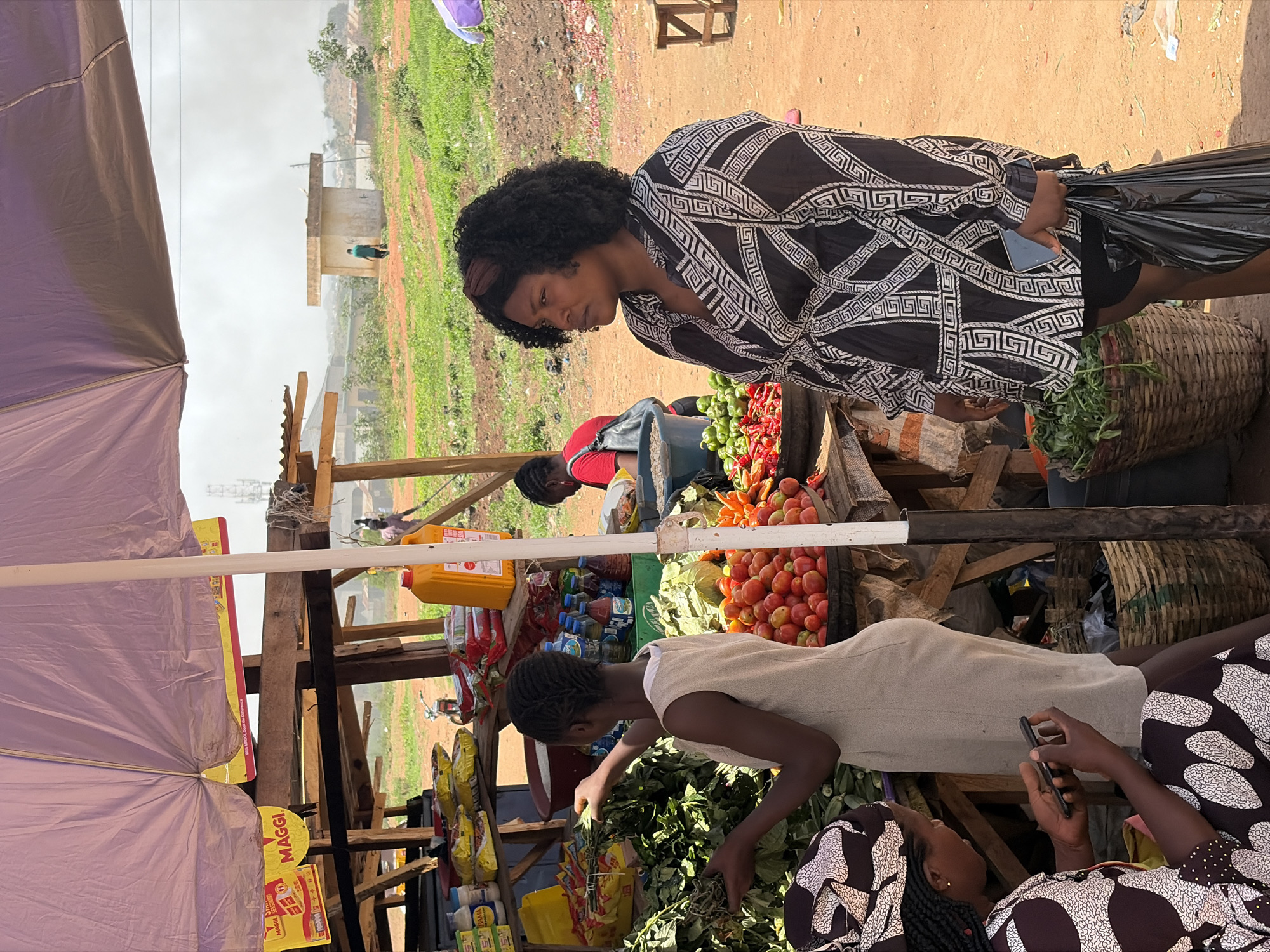 In Barakin Lerawi, Nigeria, a bustling outdoor market scene is captured. A woman with an Afro and a black and white patterned dress stands beside a market stall filled with tomatoes, green peppers, and other vegetables. She holds a black plastic bag and a mobile phone. The stall is tended by a woman in a white dress, sorting through leafy greens. Nearby, another woman, adorned in a patterned headscarf and top, is occupied with a mobile phone. A purple sunshade provides partial cover to the stall, which is constructed of wooden planks. Packets of Maggi seasoning are visible on the stall. In the background, a field stretches out, with a building and power lines visible in the distance. Other market-goers are seen indistinctly behind the stall.