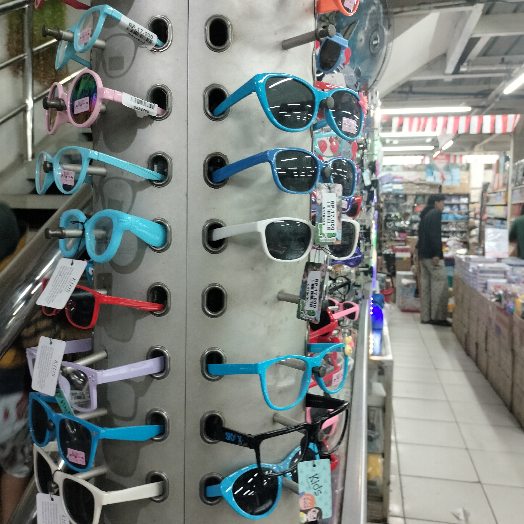 An eye-level, medium shot captures a display of colorful sunglasses for children in a retail store. The sunglasses are arranged vertically on a grey metal rack, each held in place by a cylindrical protrusion. From top to bottom, the rack displays: light blue glasses with a price tag that reads "RP. 17.000", pink framed glasses with reflective lenses and a price tag, and more light blue glasses. Below these, a row of darker blue framed glasses with polarized lenses is visible, followed by white framed glasses with dark lenses and a price tag. Further down are purple framed glasses with a "KIDS" tag, bright blue framed glasses, black framed glasses with "SKY X" printed on the arm, and another pair of blue framed glasses with a "Kids" tag featuring cartoon characters. The background shows aisles of shelves filled with merchandise, a person in a black jacket and light-colored pants walking away from the camera, and white and red striped awnings. The floor is tiled. The scene is illuminated by overhead fluorescent lighting. The store appears to be located in South Jakarta, Indonesia, as suggested by the price format on the tags.