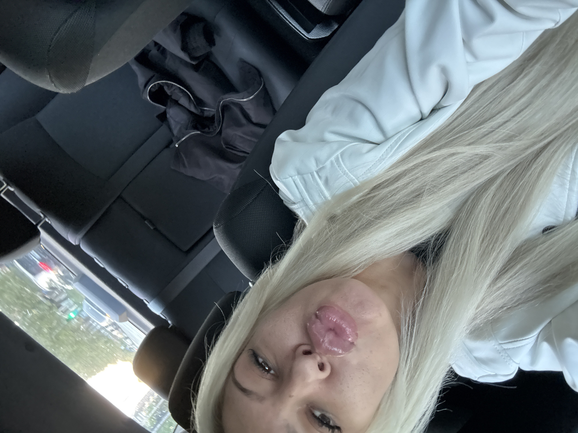 An overhead, slightly rotated shot captures a woman inside a car, looking directly at the camera with a playful expression. Her long, straight blonde hair cascades over her shoulder and chest, and she is wearing a white, possibly leather or faux-leather, jacket. Her tongue is partially extended, touching the area between her upper lip and cheek. Her eyes are dark, and she has visible freckles on her face.
The setting is the interior of a vehicle, likely a car, during the daytime. The woman appears to be seated in the front passenger seat or possibly the back. A black jacket is visible crumpled on an adjacent seat or surface, contrasting with the woman's light-colored attire. The car's dark interior, including headrests and seat fabric, is visible around her.
Through the window in the background, a blurry outdoor scene is discernible, showing greenery, parts of buildings, and what appears to be traffic lights (red and green blurs), suggesting an urban or suburban environment consistent with Plovdiv, Bulgaria. The natural light indicates it is daytime. No discernible text is visible in the image.