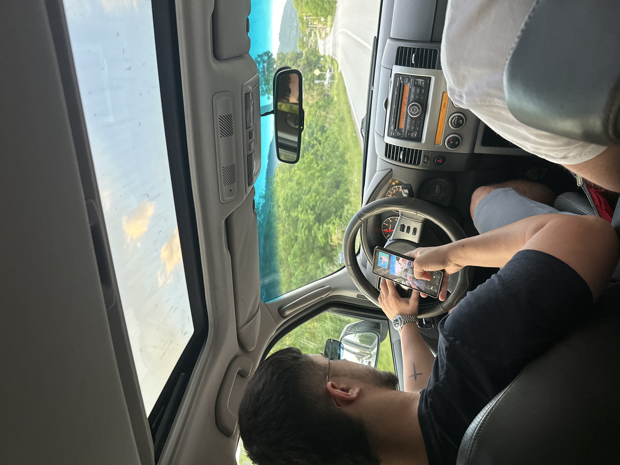 The image shows the interior of a car from a low angle, looking up and forward. A man is in the driver's seat, holding a smartphone in his hands. He has dark hair, a beard, and is wearing glasses and a watch. He has a cross tattoo on his left arm. The phone screen is visible, showing several images.
The car's dashboard, steering wheel, and rearview mirror are visible, as well as a partial view of the passenger's seat, occupied by a person wearing a white shirt. The car has a sunroof.
Through the windshield and sunroof, a green, wooded landscape and a road are visible.
Location: Unknown, Unknown.